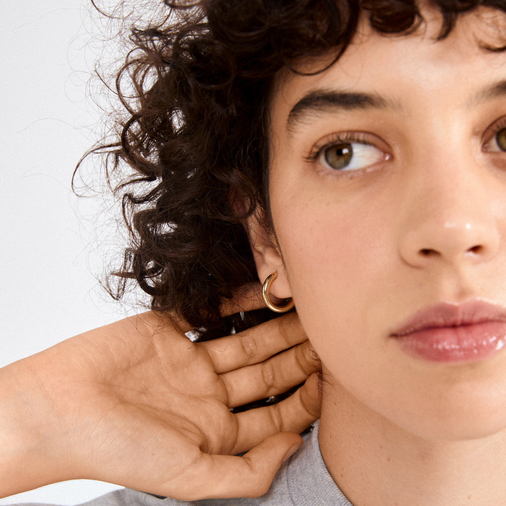 Person with dark curly hair wearing DOLAG gold-plated chunky hoop earring, hand behind ear, showcasing the earring's bold, stylish design.