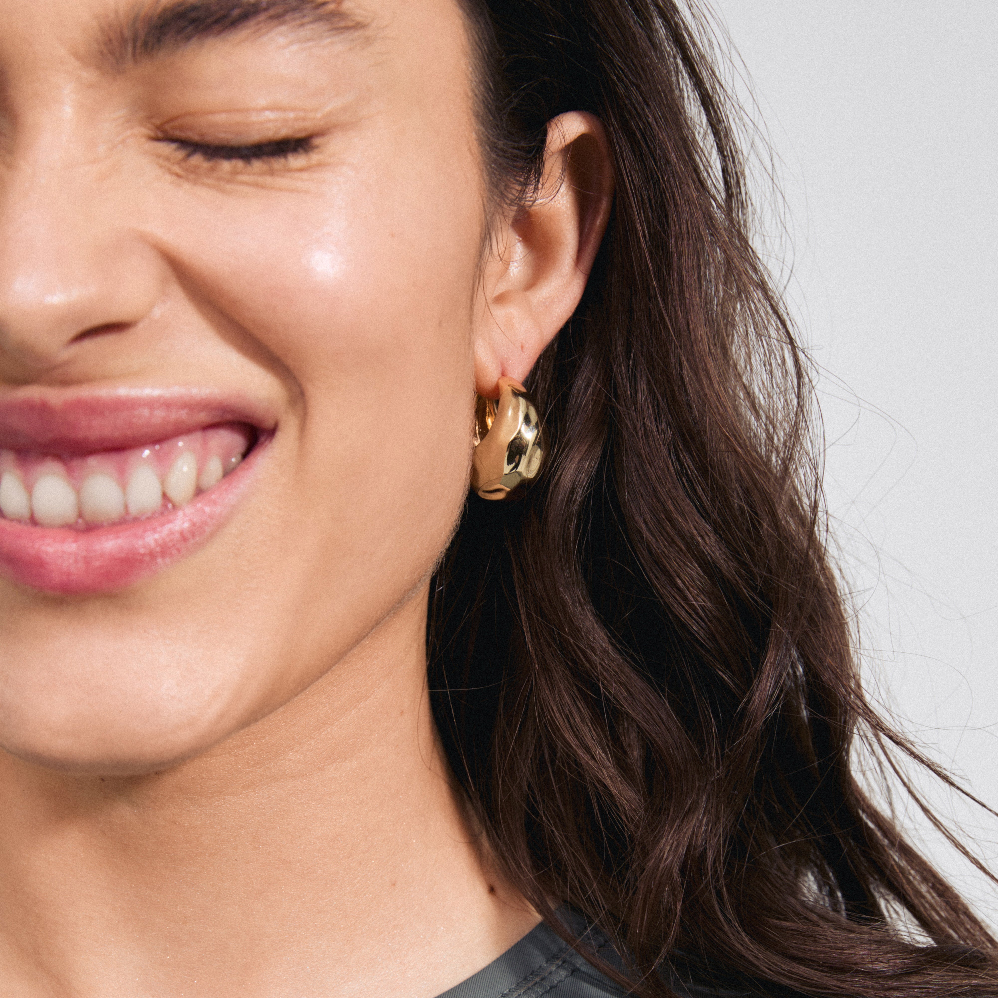 Woman wearing BELIEVE chunky hoop earrings gold-plated with a shiny, hammered texture, smiling, showcasing the earrings' light-catching design and easy-to-use click clasps.