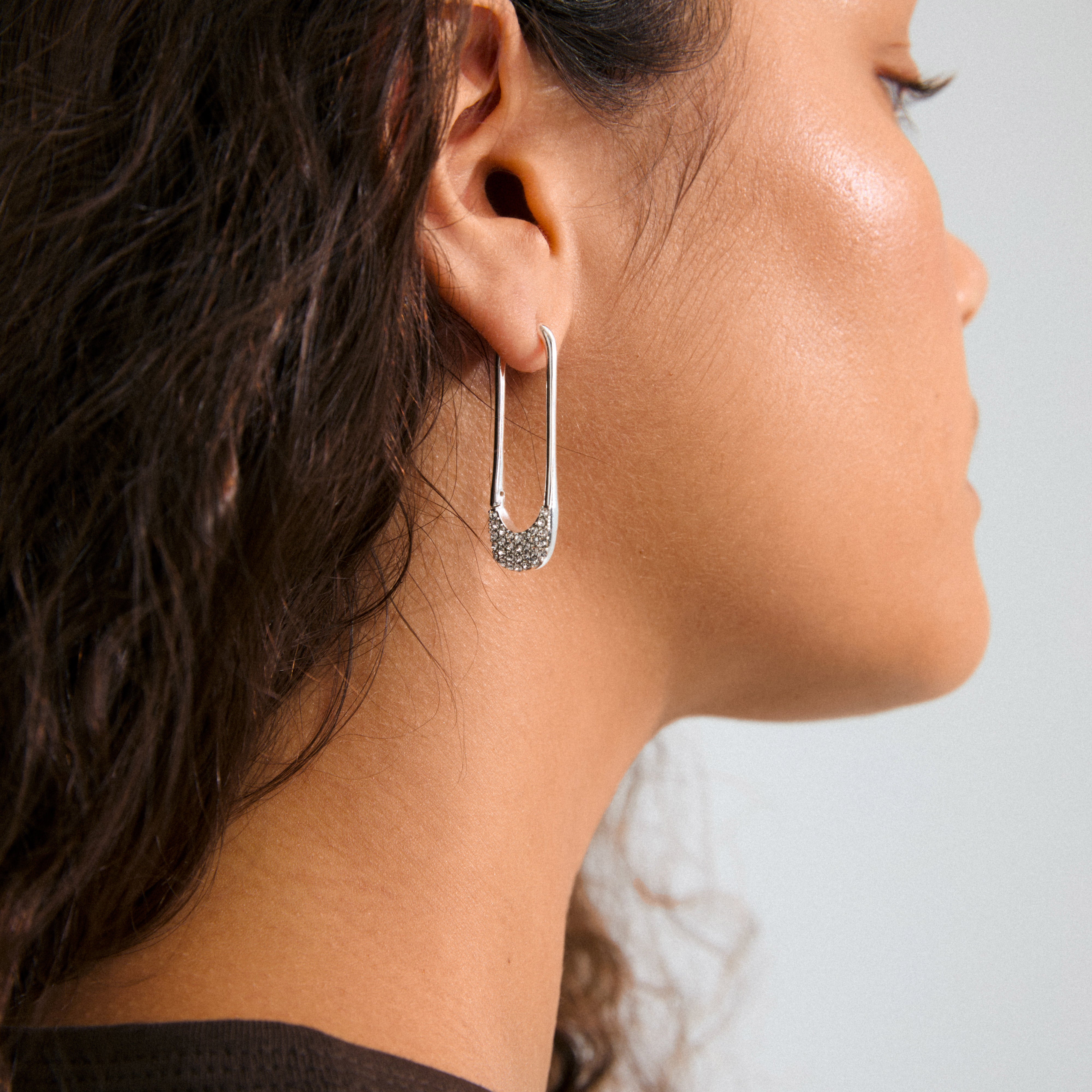 Side profile of a woman showcasing Pilgrim's INSTANT hoops, silver-plated earrings inspired by safety pins, adorned with sparkling crystals, emphasizing edgy yet sophisticated style.