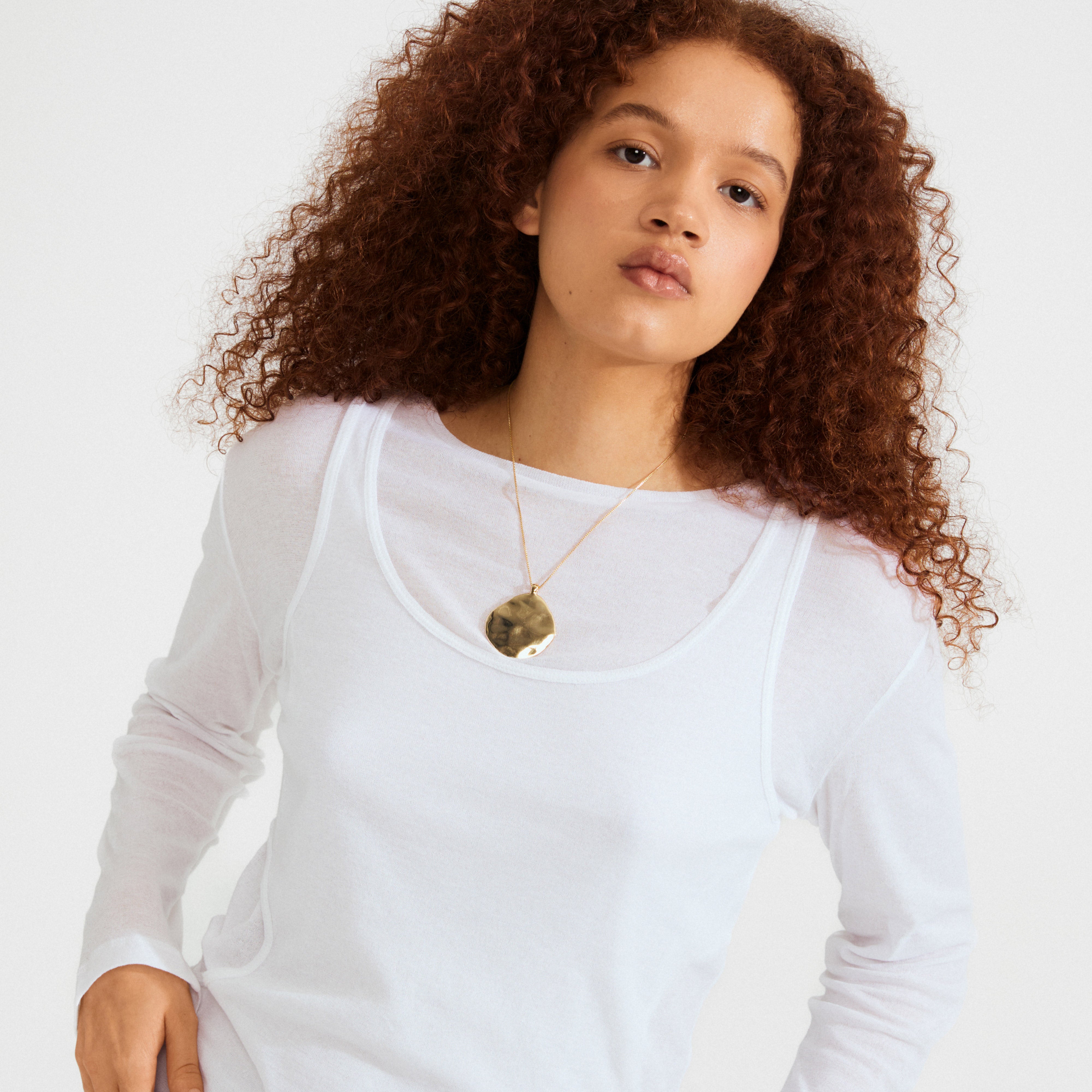 Young person wearing Pilgrim's SPARKLE necklace: a gold-plated chain with a large, wavy hammered pendant, styled over a layered white top.