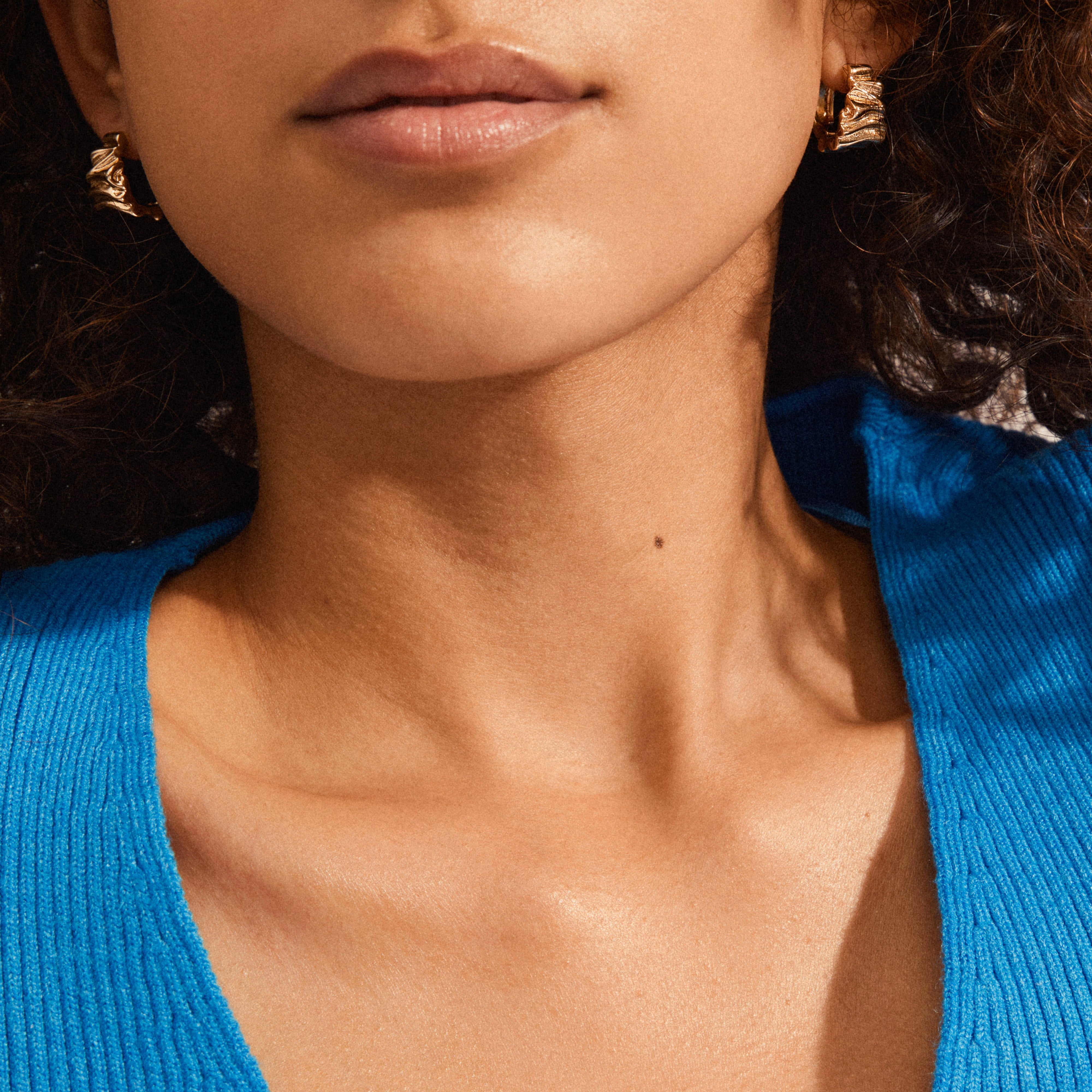 WILLPOWER huggie hoop earrings gold-plated, showcasing Nordic design with organic-embossed texture, worn by a model highlighting natural skin texture and a ribbed blue top.