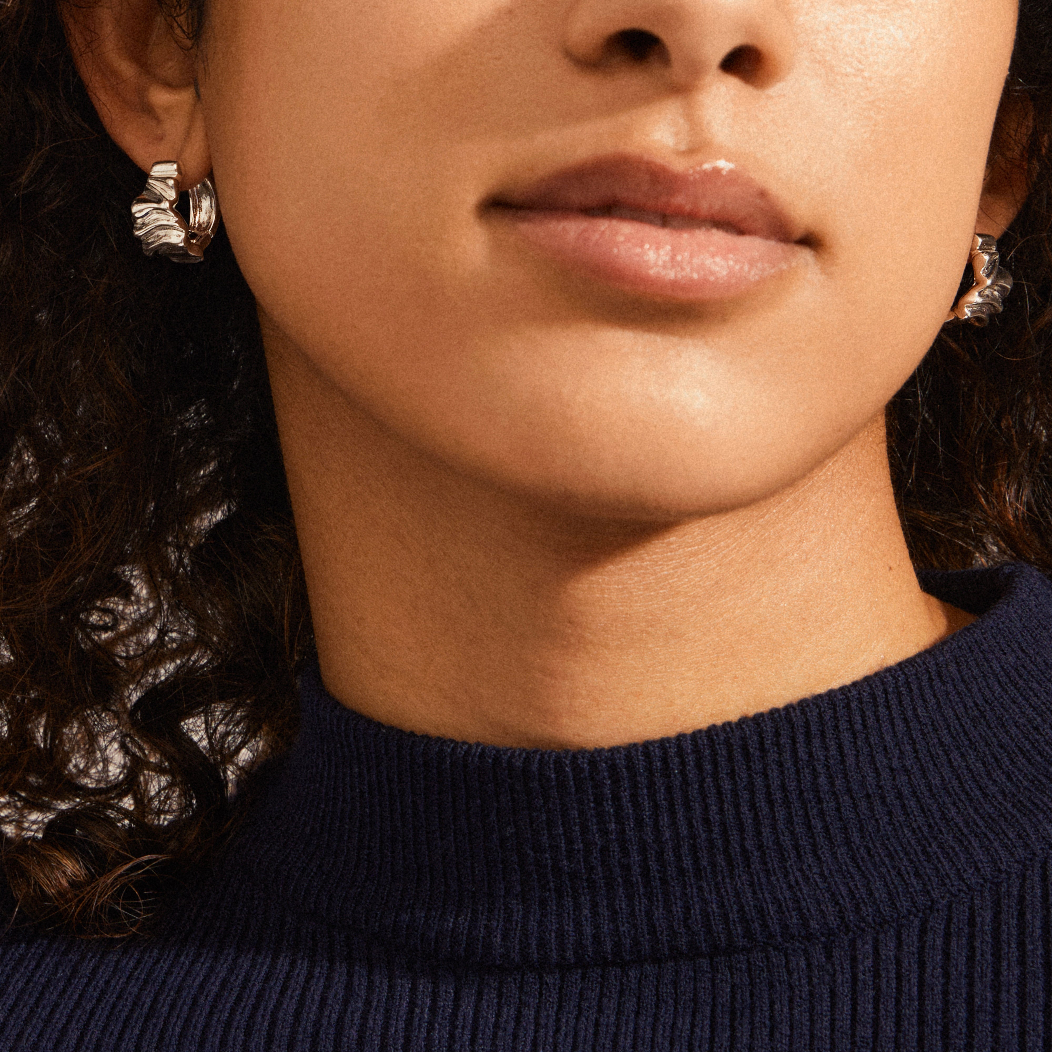 Close-up of a person wearing Pilgrim's WILLPOWER silver-plated huggie hoop earrings, showcasing their wavy, handcrafted design against a navy ribbed-knit turtleneck.