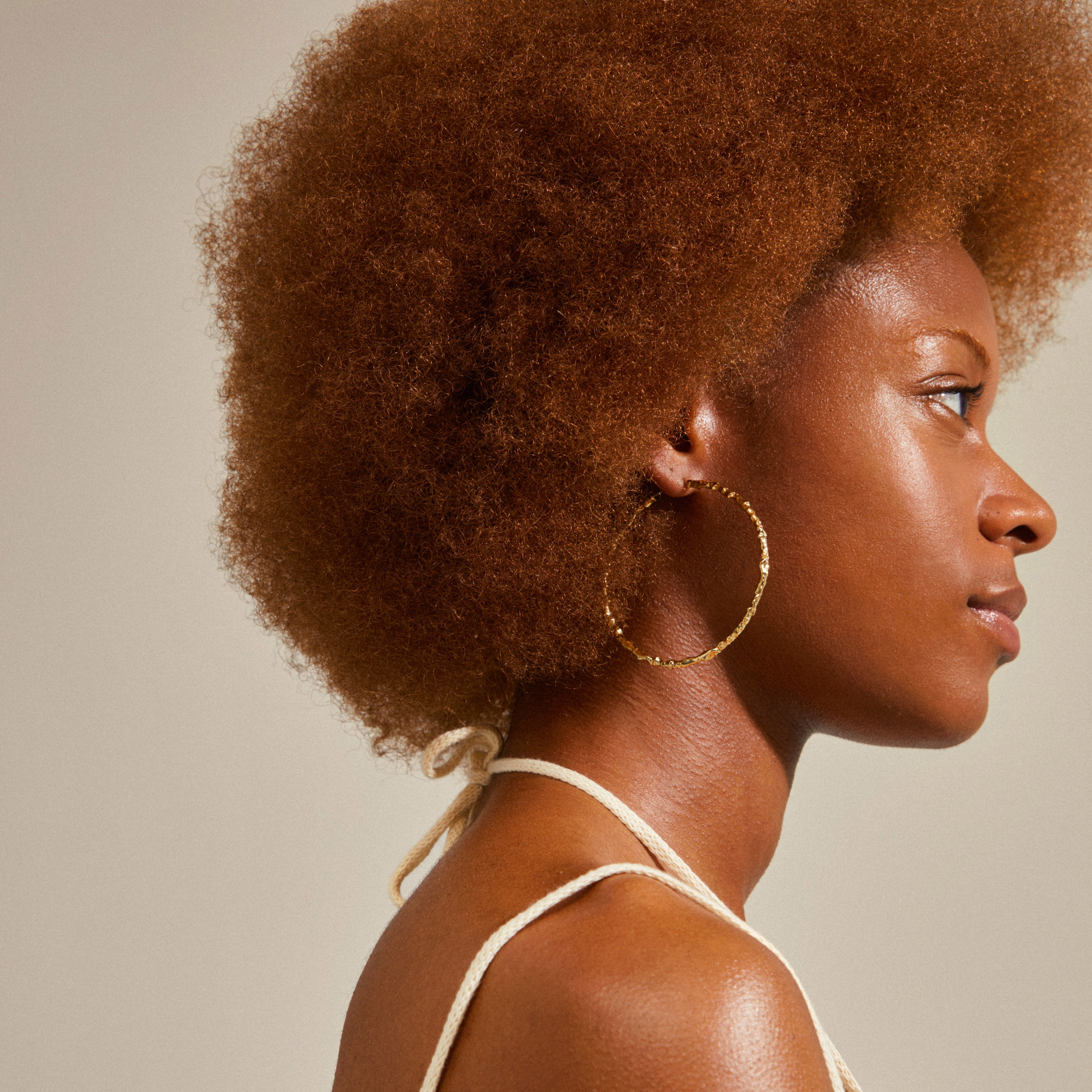 SUN mega hoops gold-plated earrings worn by a woman with curly hair, showcasing their large, uneven design, embodying elegance and versatility for summer adventures.