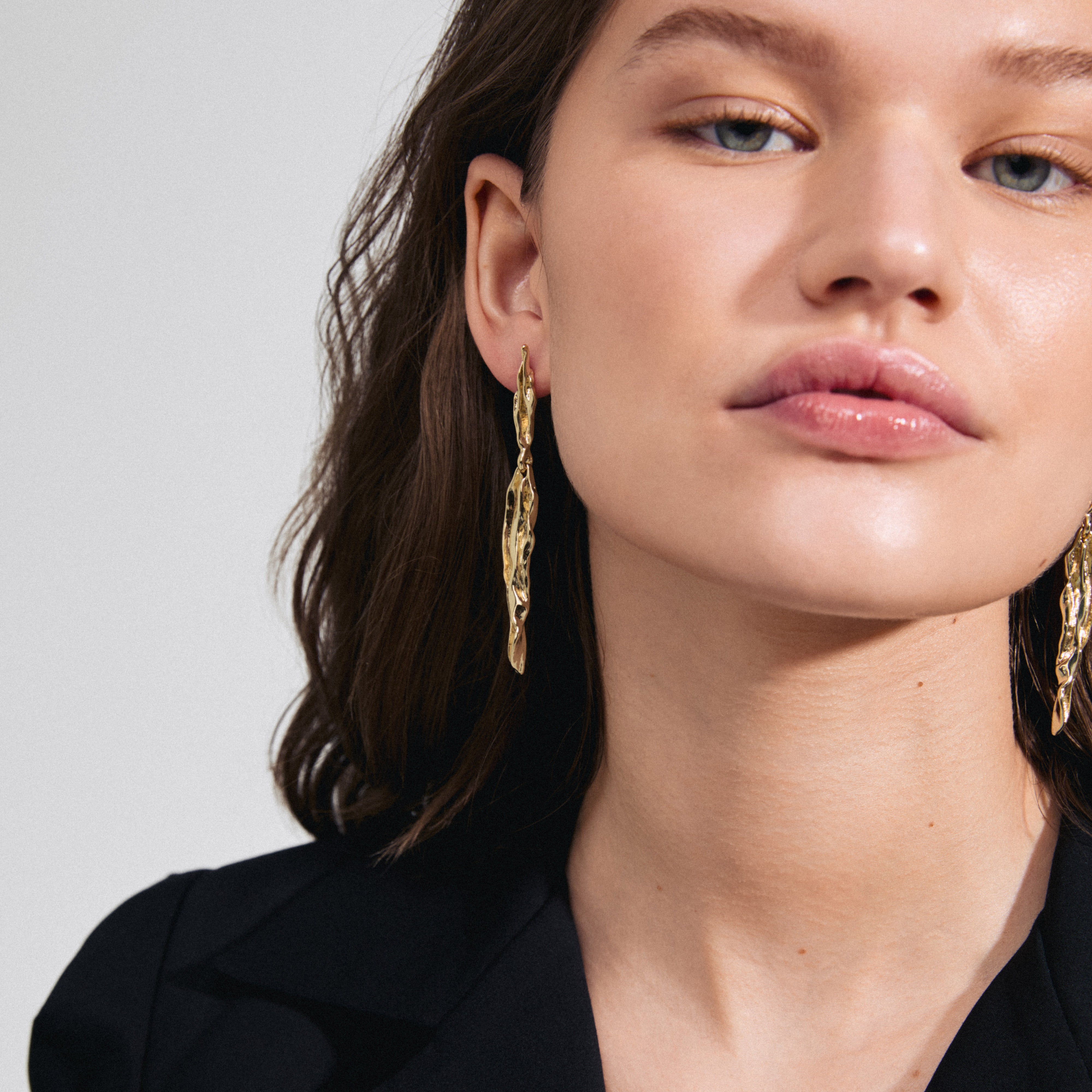 FEEL earrings gold-plated from Pilgrim, worn by a woman, showcasing their sleek, wavy design inspired by nature, adding a luxe touch to her style.