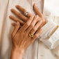 Hands overlapping on cream fabric, each adorned with the Pilgrim UNFOLD ring in gold-plated, showcasing a knotted, sculptural design with a luxurious finish.