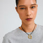 Young woman models Pilgrim's UNFOLD necklace, featuring gold-plated interlocking loops on a black cord, paired with a light-gray T-shirt.