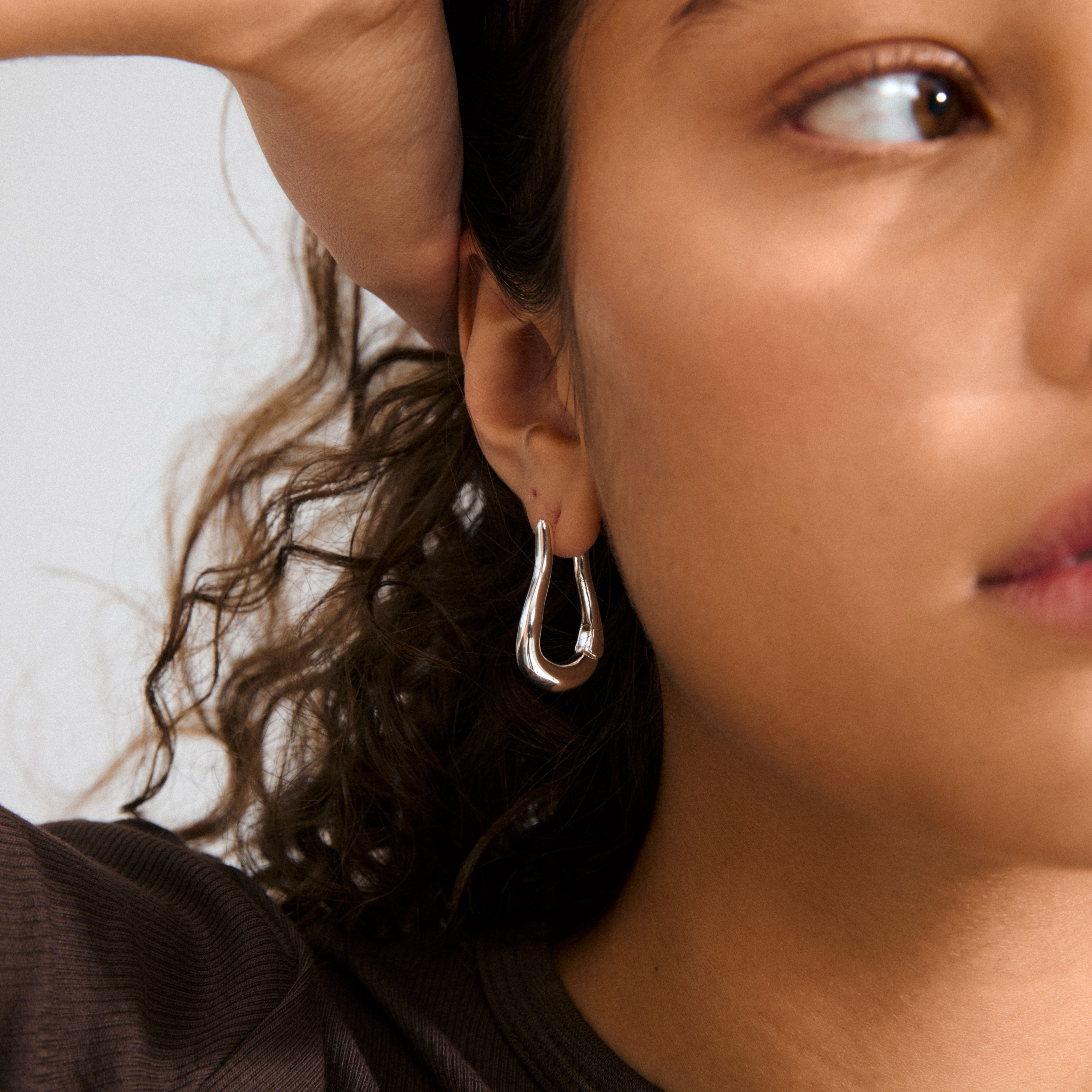 Close-up of a person's ear wearing the silver-plated UNFOLD hoops by Pilgrim, featuring a free-form design and shiny finish, complementing dark curly hair.