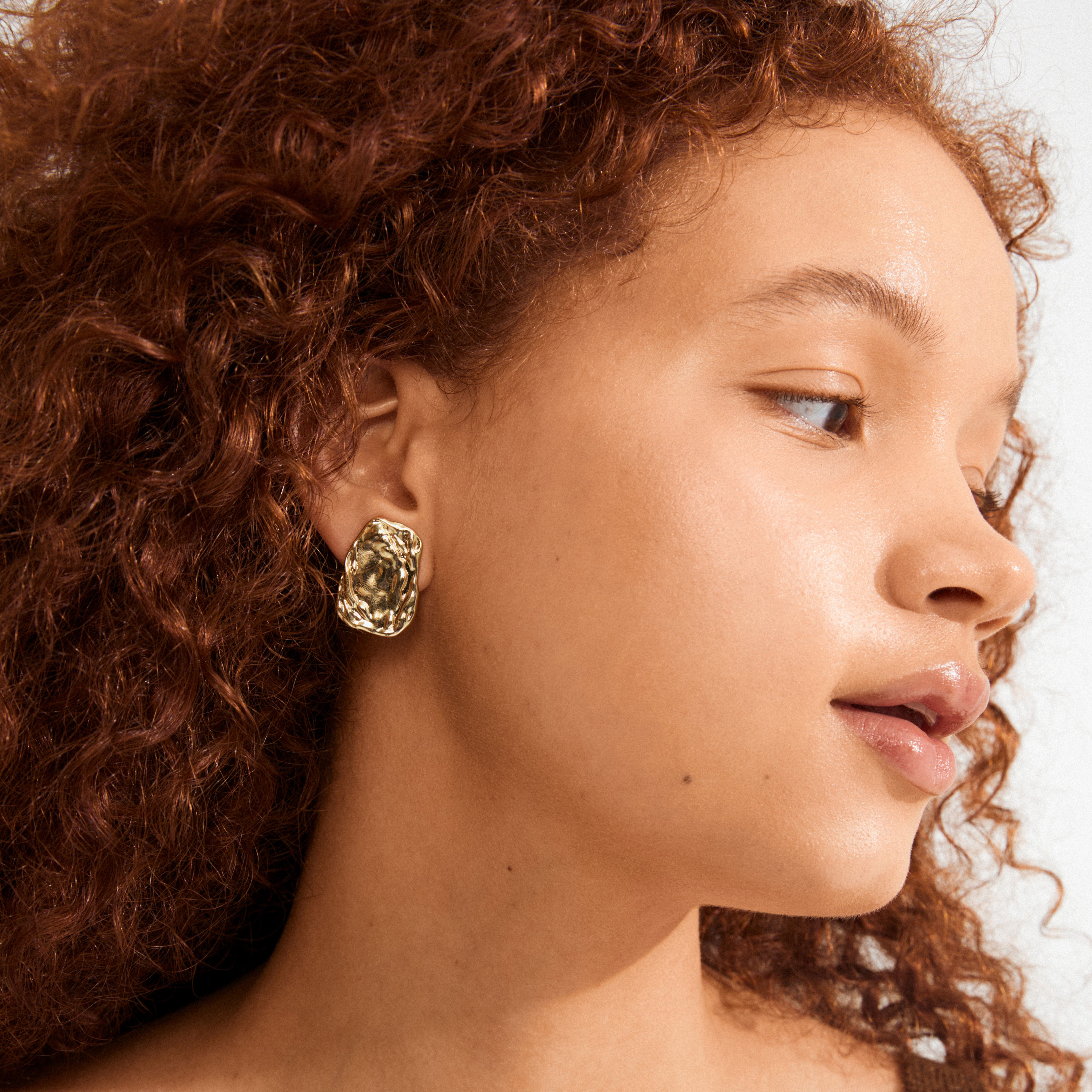 Profile view showcasing a young person wearing the gold-plated SHIFT earring by Pilgrim, highlighting its chunky, hammered-metal design and handcrafted finish.