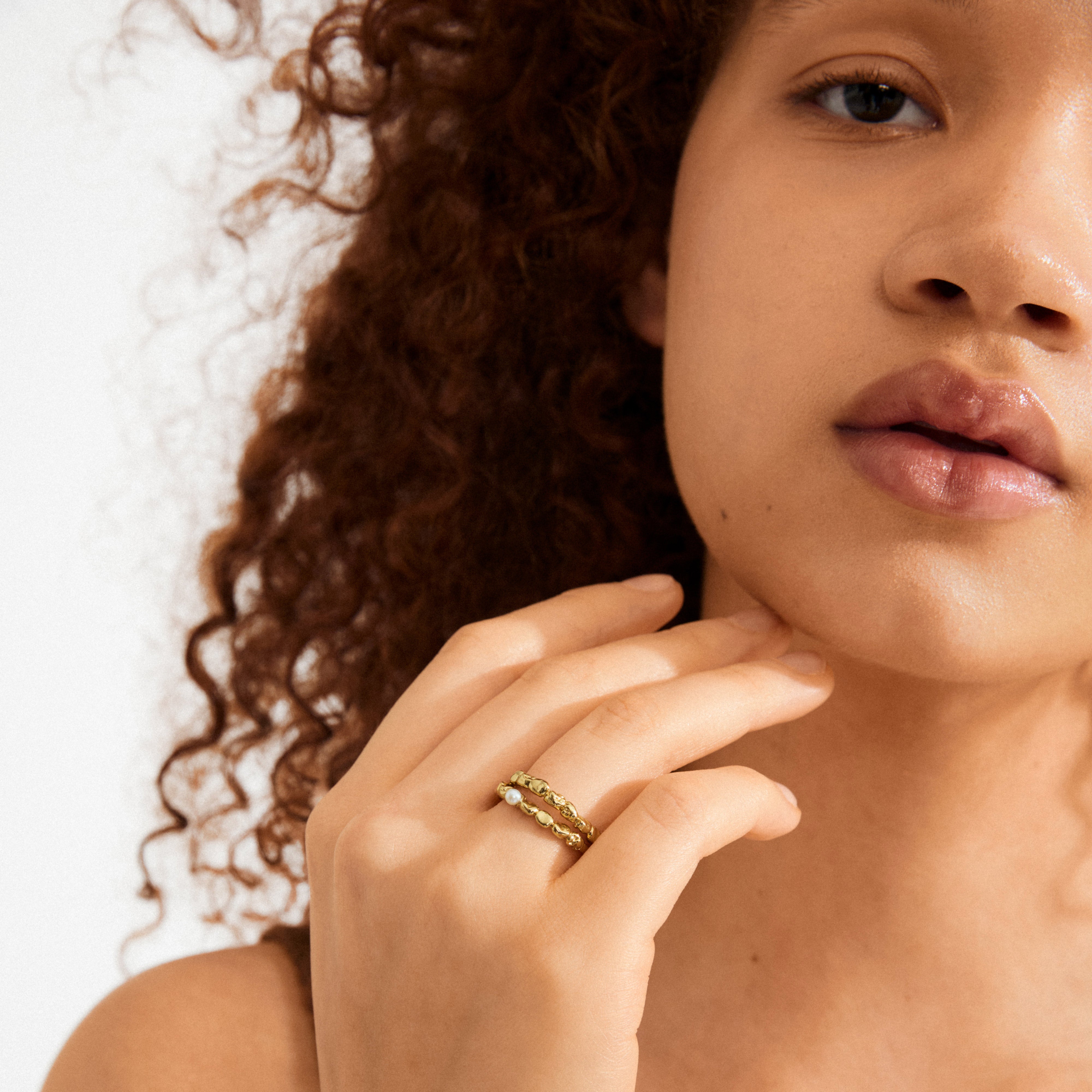 Young person with curly hair models Pilgrim's SHIFT ring gold-plated set, featuring textured bands and a pearl accent, hand poised under chin.