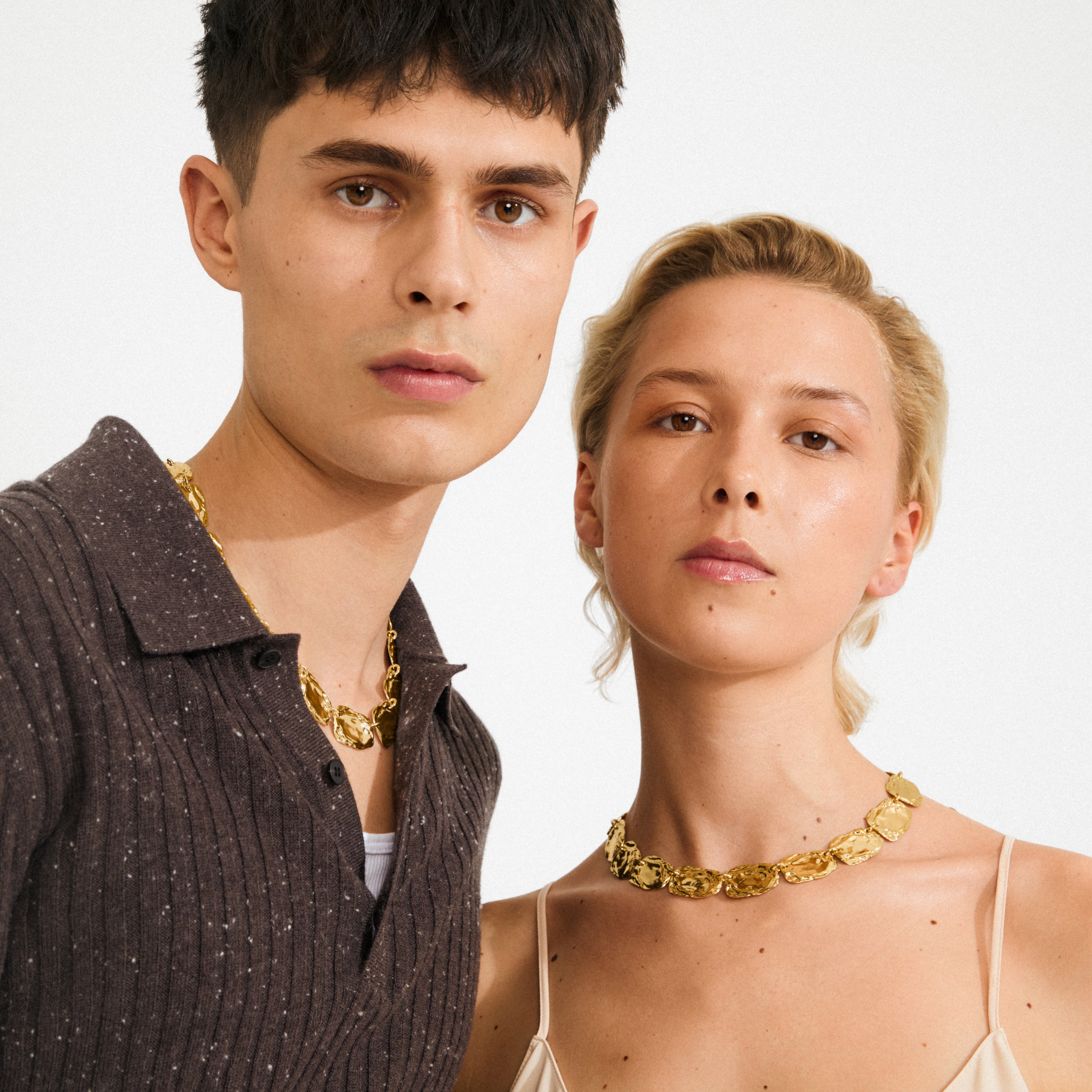 Two young adults wearing Pilgrim's SHIFT gold-plated necklaces, showcasing chunky, rustic coin links as bold statement pieces for diverse styling.