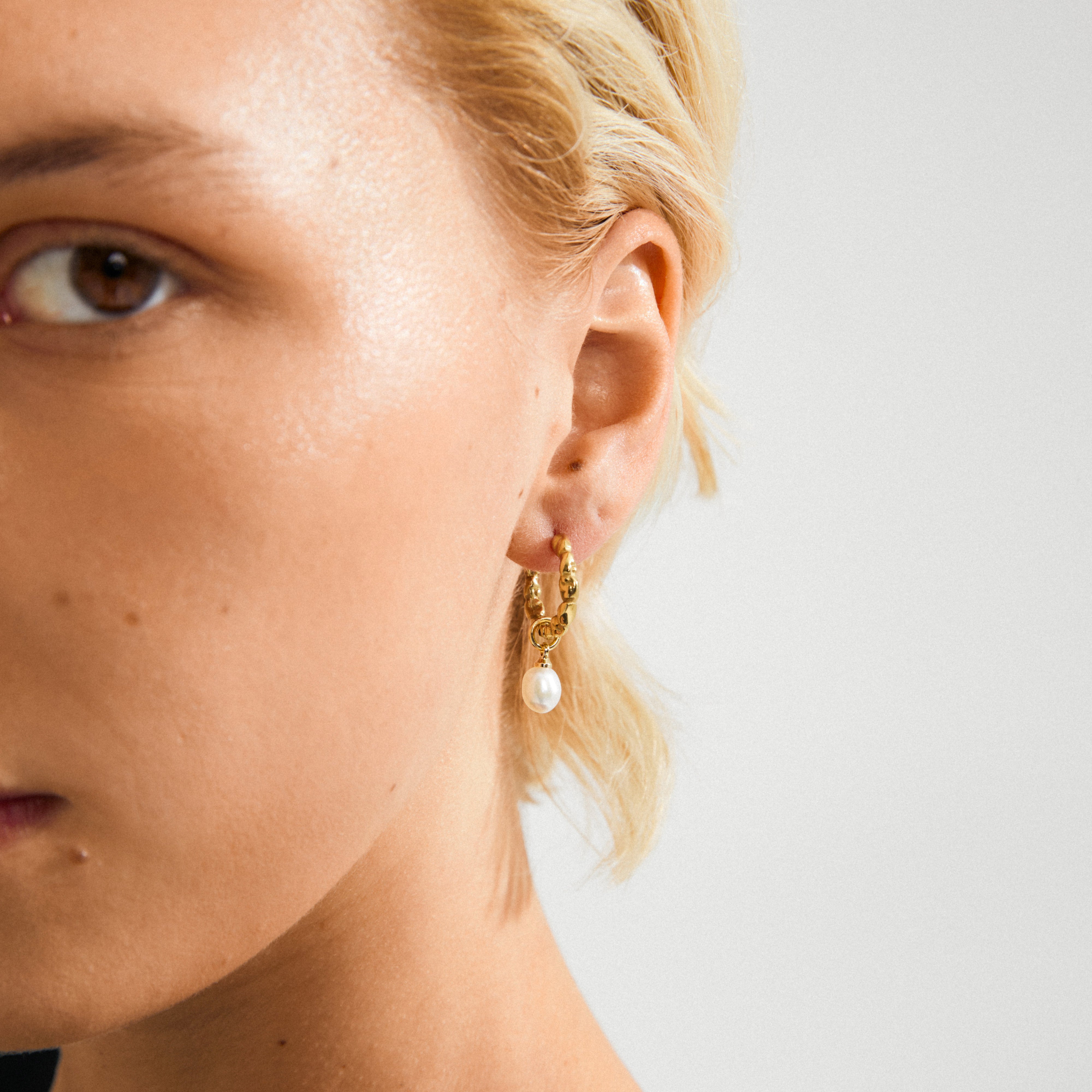 Gold-plated SHIFT pearl earrings featuring a small textured hoop with a removable white freshwater pearl, shown on a person with light-blonde hair.