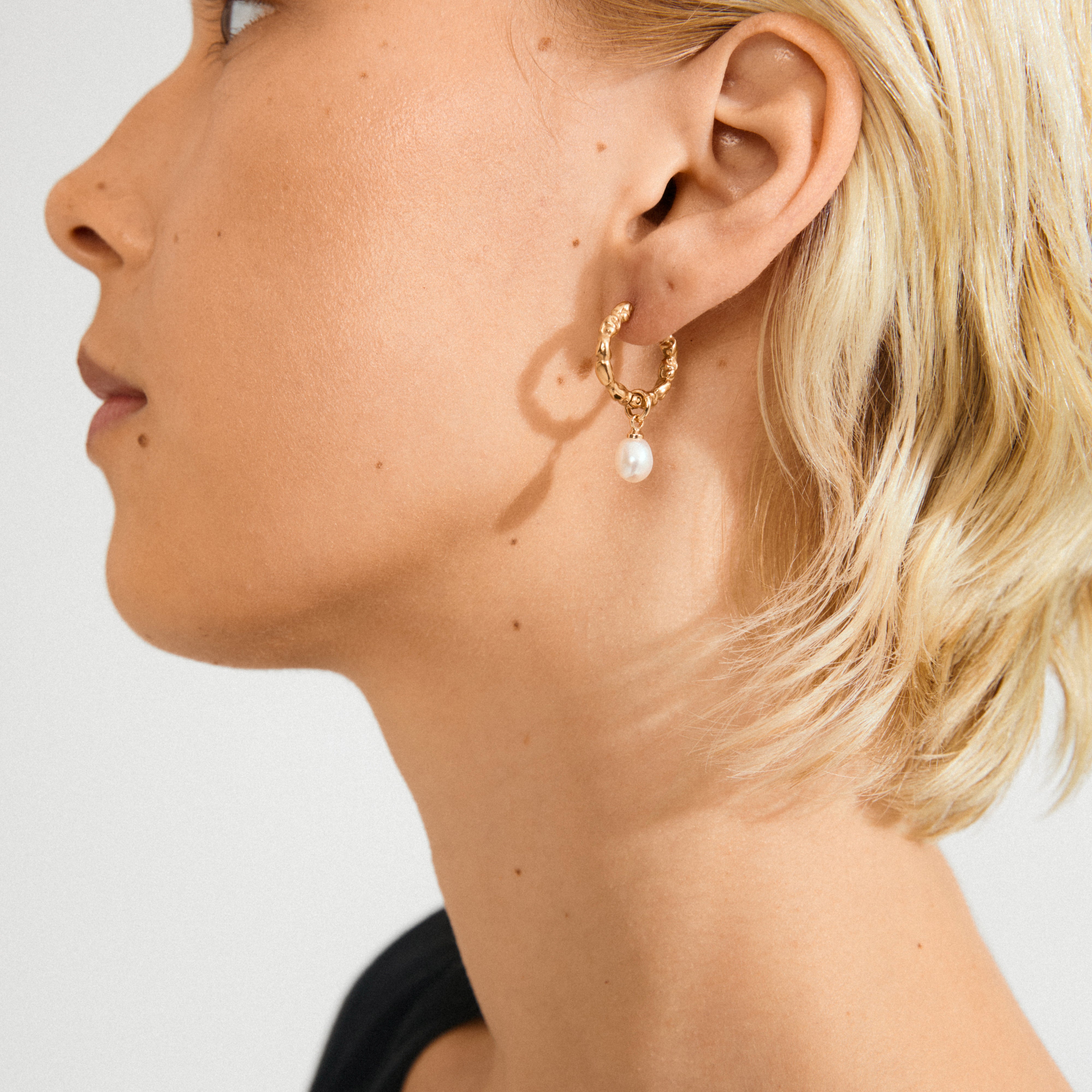 Close-up of a person wearing Pilgrim's SHIFT gold-plated hoop earrings with a dangling freshwater pearl, showcasing the earring's shiny finish and textured detail.