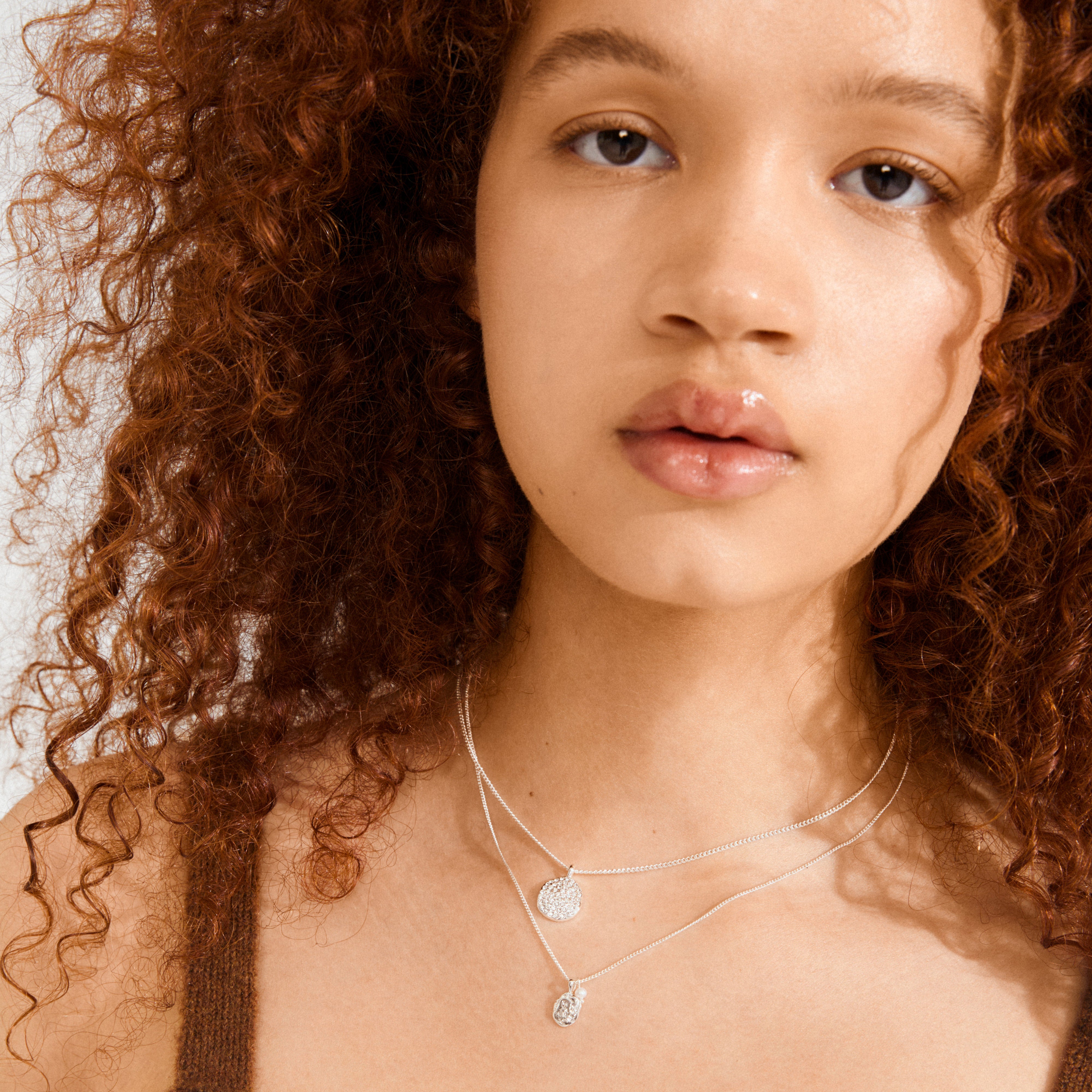 Young person with curly auburn hair wears Pilgrim's SHIFT necklace 2-in-1 set, featuring layered silver chains with crystal and pearl pendants, exuding a boho-chic vibe.