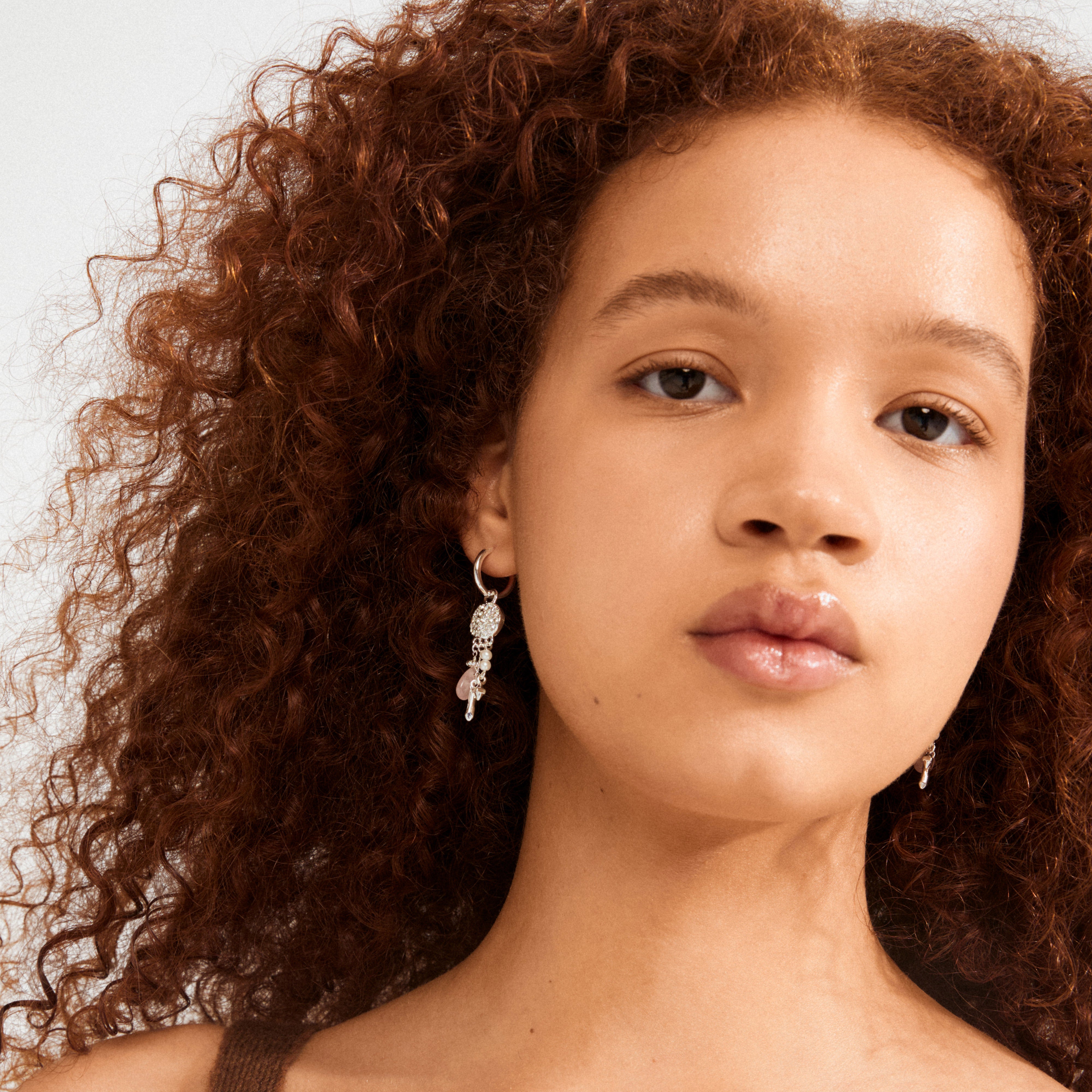Young person with curly auburn hair wearing Pilgrim's SHIFT hoop earrings, featuring silver charms, Preciosa crystals, and rose quartz, exuding a whimsical, dreamy vibe.