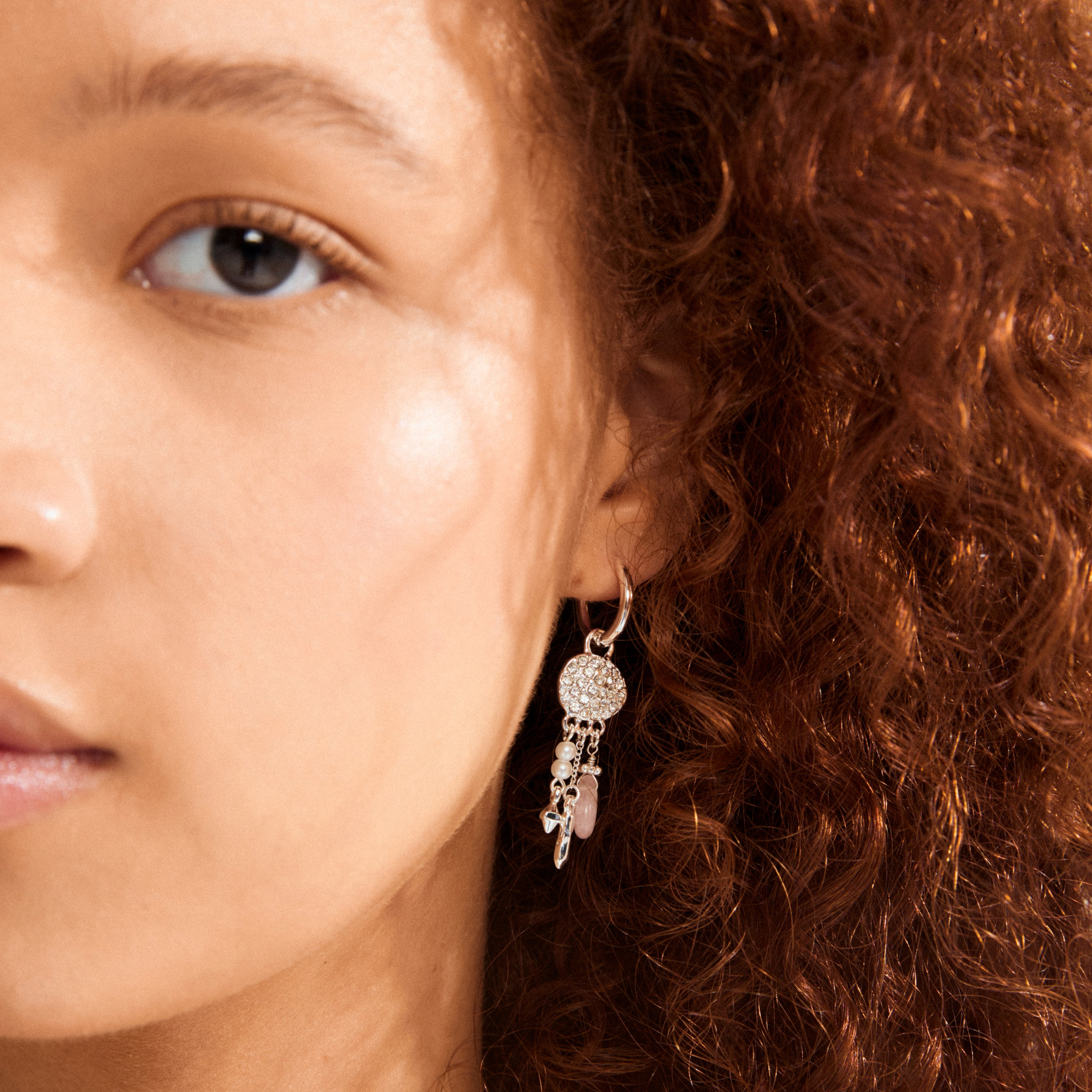 SHIFT hoop earrings, silver-plated, featuring crystal-studded charms with dangling pearls and beads, showcased on a person's cheek and auburn hair.