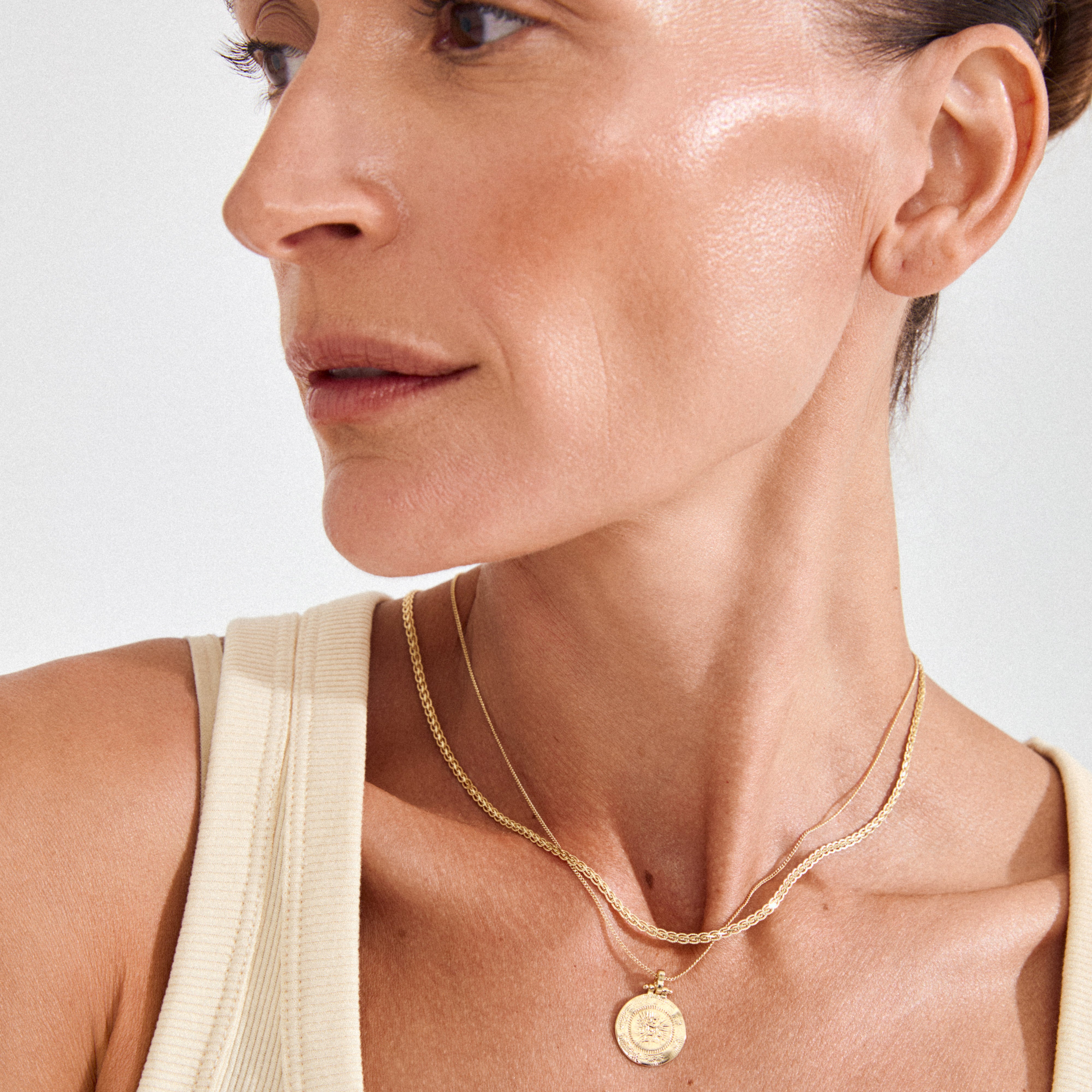 NOMAD 2-in-1 coin necklace gold-plated, featuring a sleek curb chain and medallion pendant, layered around a cream rib-knit tank top.