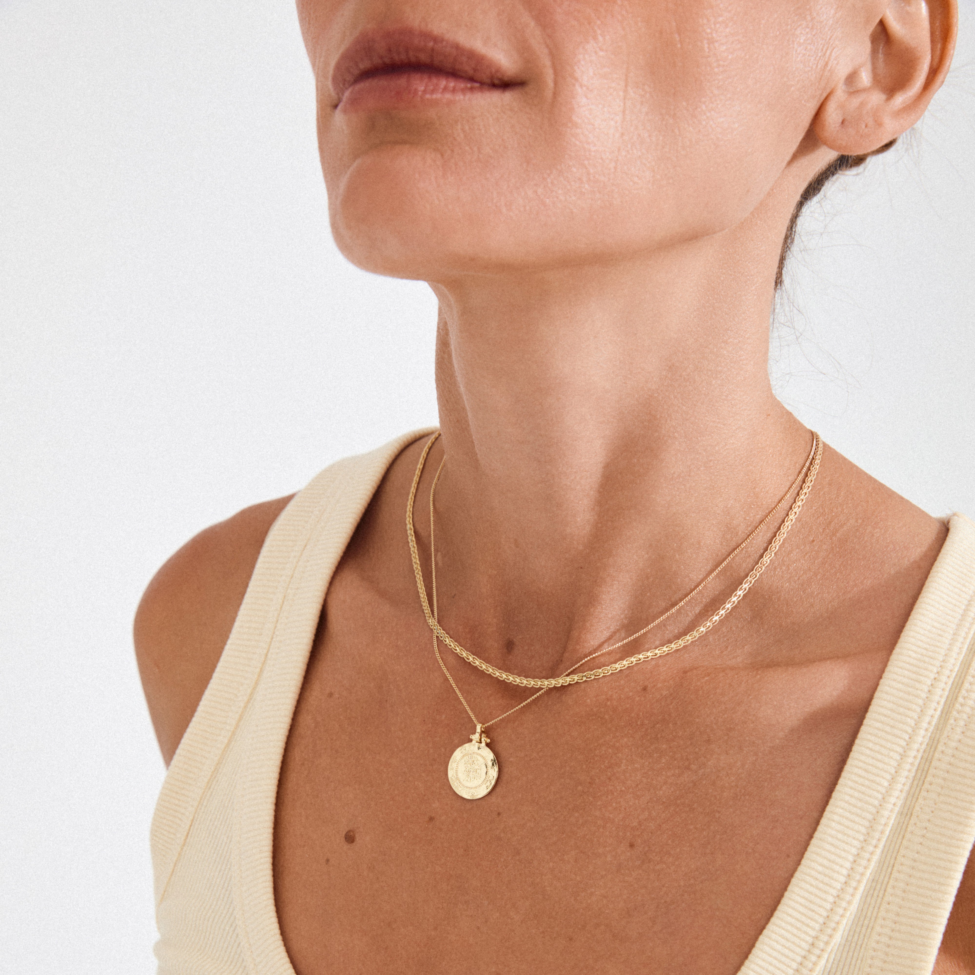 Close-up of the NOMAD 2-in-1 coin necklace gold-plated, featuring a curb chain and a coin pendant chain, worn on a ribbed tank top.