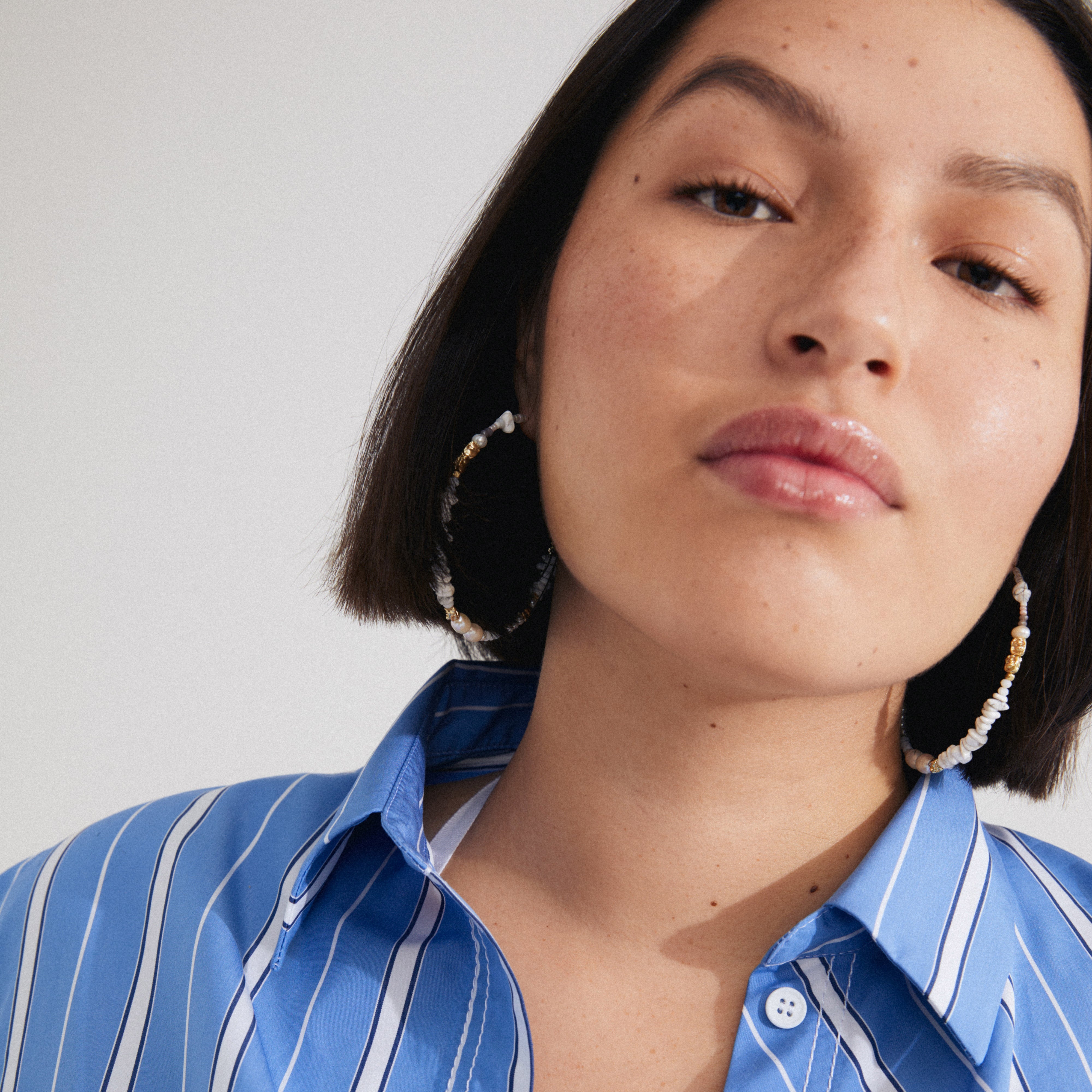 FORCE mega hoop earrings in white and gold-plated, worn by a woman with short dark hair, complementing her blue striped shirt and casual summer style.