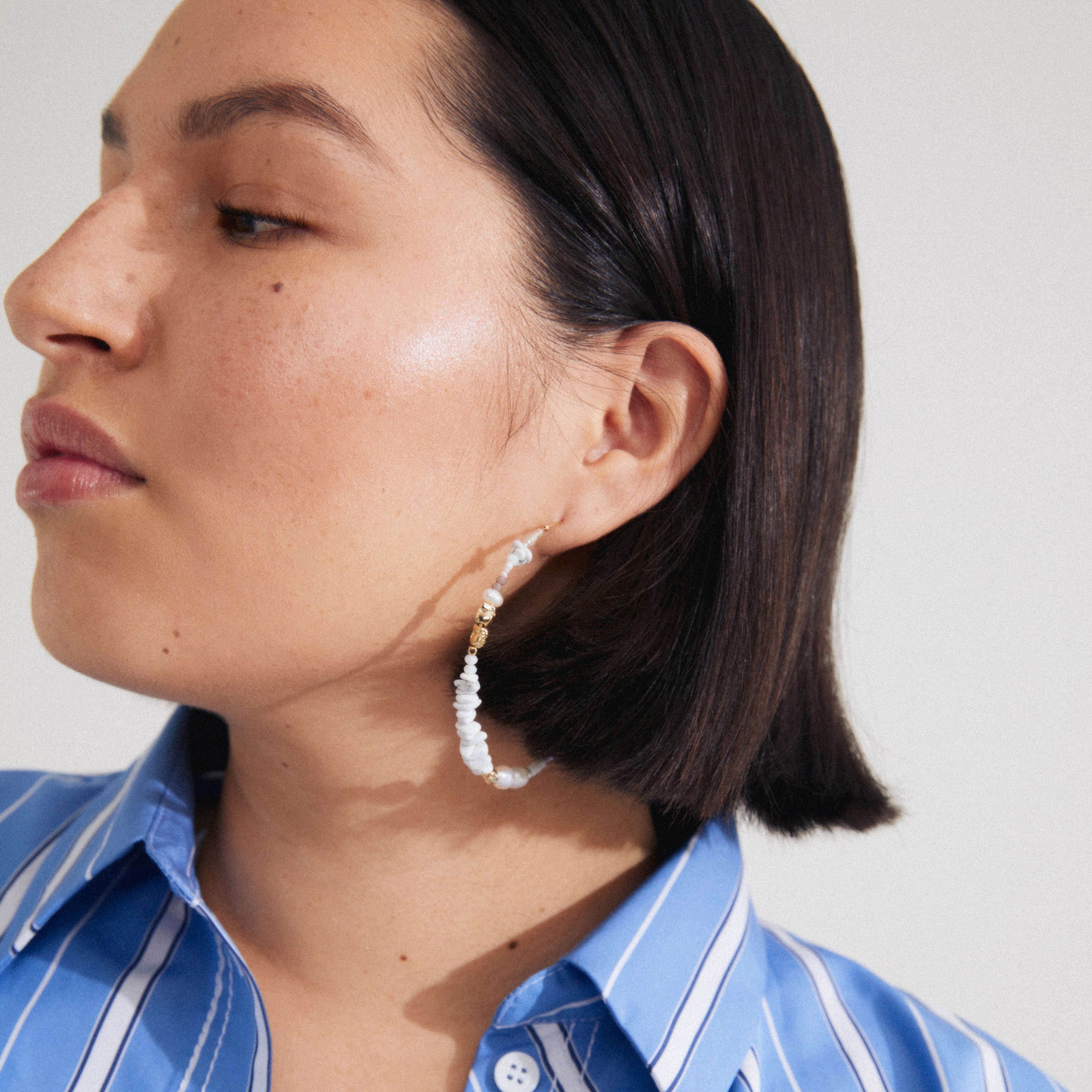 FORCE mega hoop earrings in white and gold-plated, featuring white turquoise and freshwater pearls, worn by a woman in a blue striped shirt.