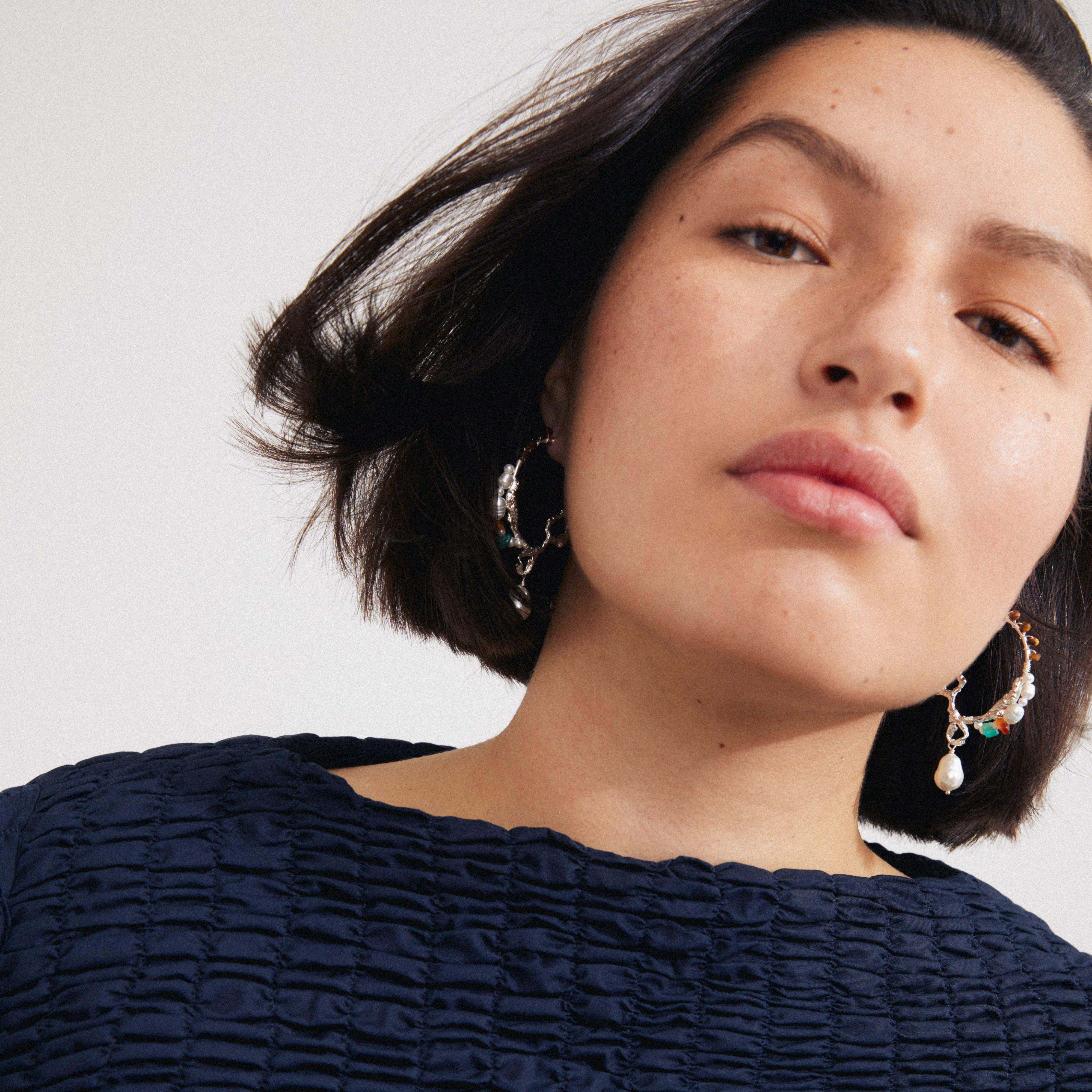Woman with short dark hair wearing Pilgrim's FORCE large hoop earrings, featuring freshwater pearls and semi-precious gemstones, showcasing an organic, bohemian silver-plated design.