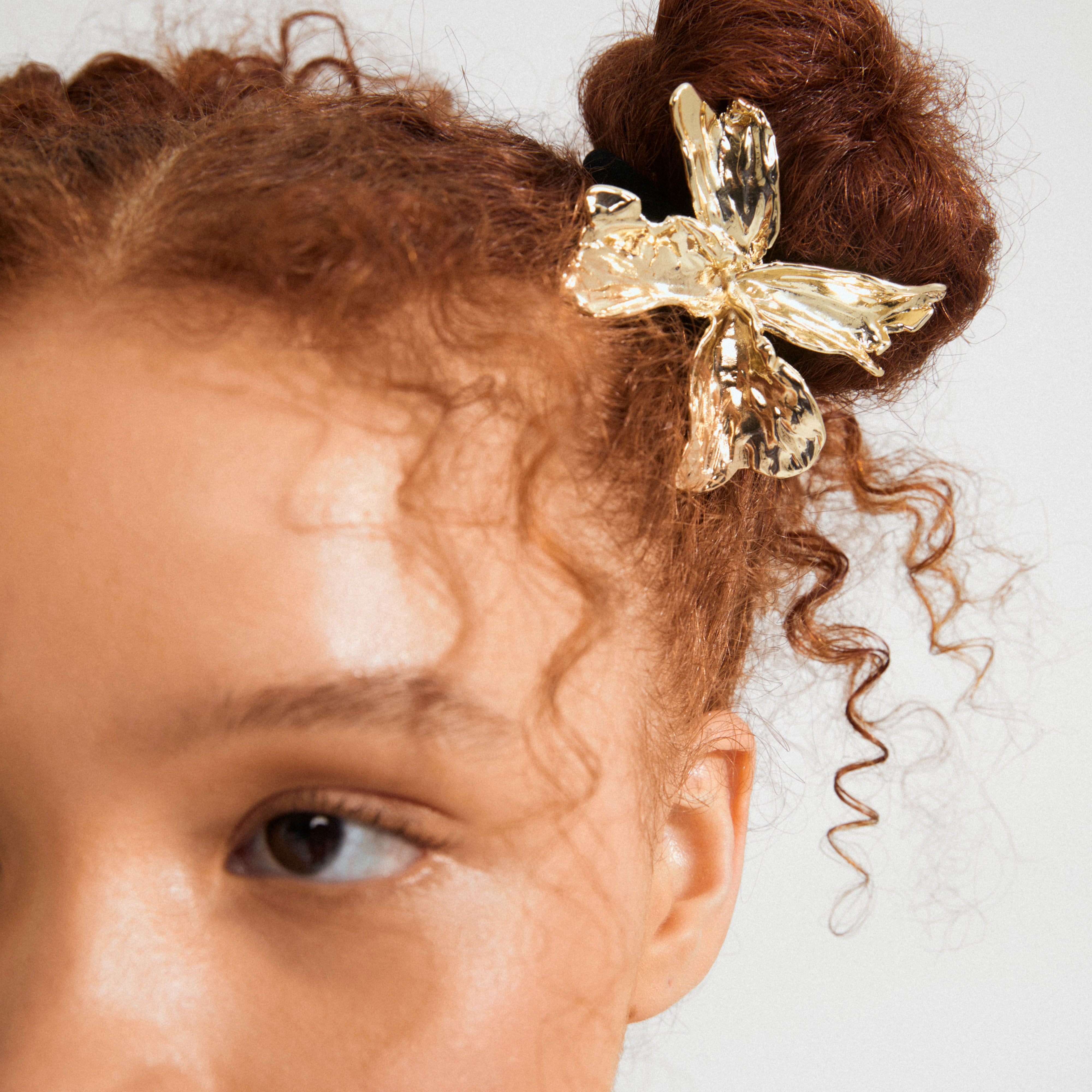 Close-up of a bun adorned with Pilgrim's DREAM hair-accessory, a gold-plated flower/bow ornament, enhancing curls with an elegant summer touch.