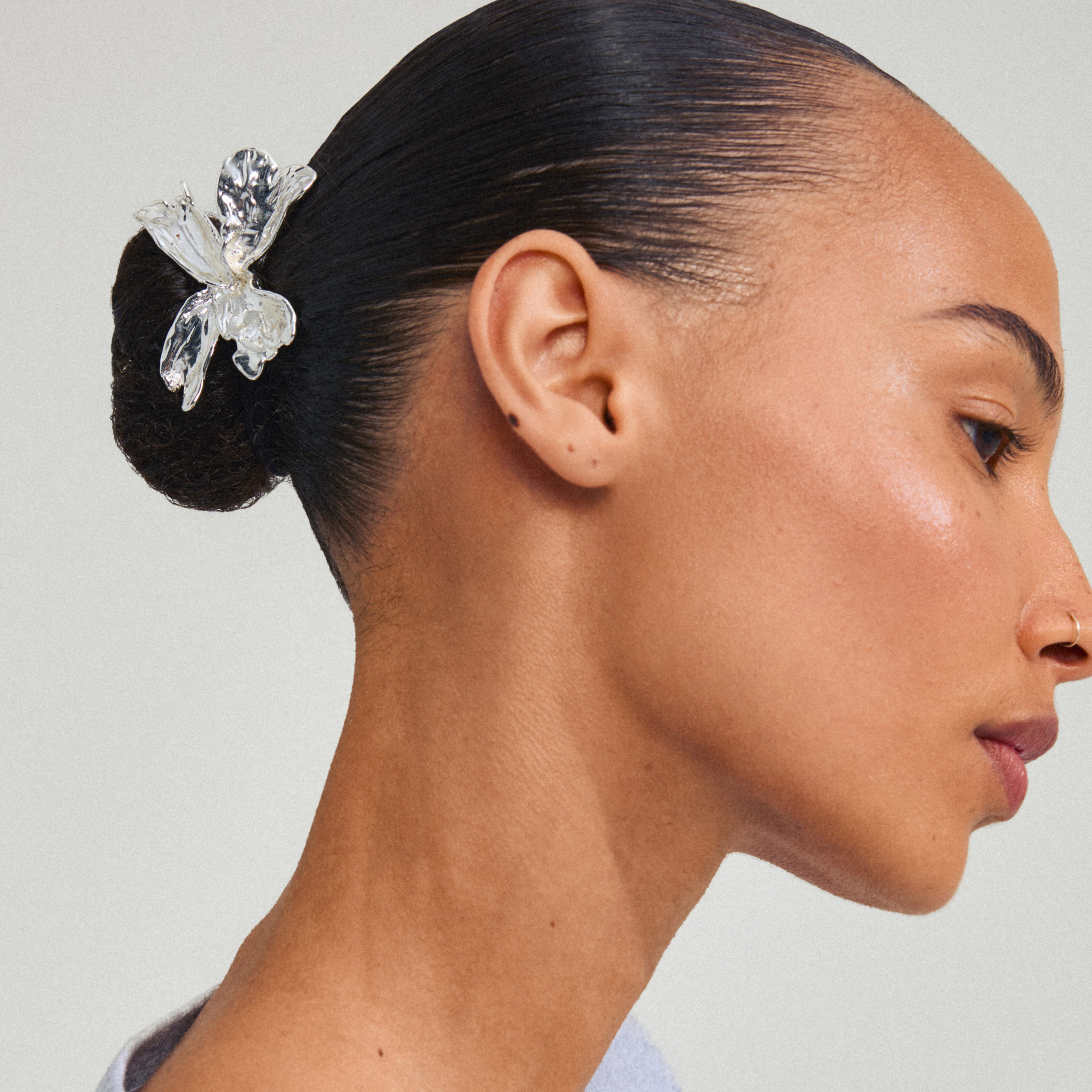 Side profile showcasing the DREAM hair-accessory, a silver-plated orchid-inspired charm securing a neat bun, complemented by minimal earrings and a nose ring.