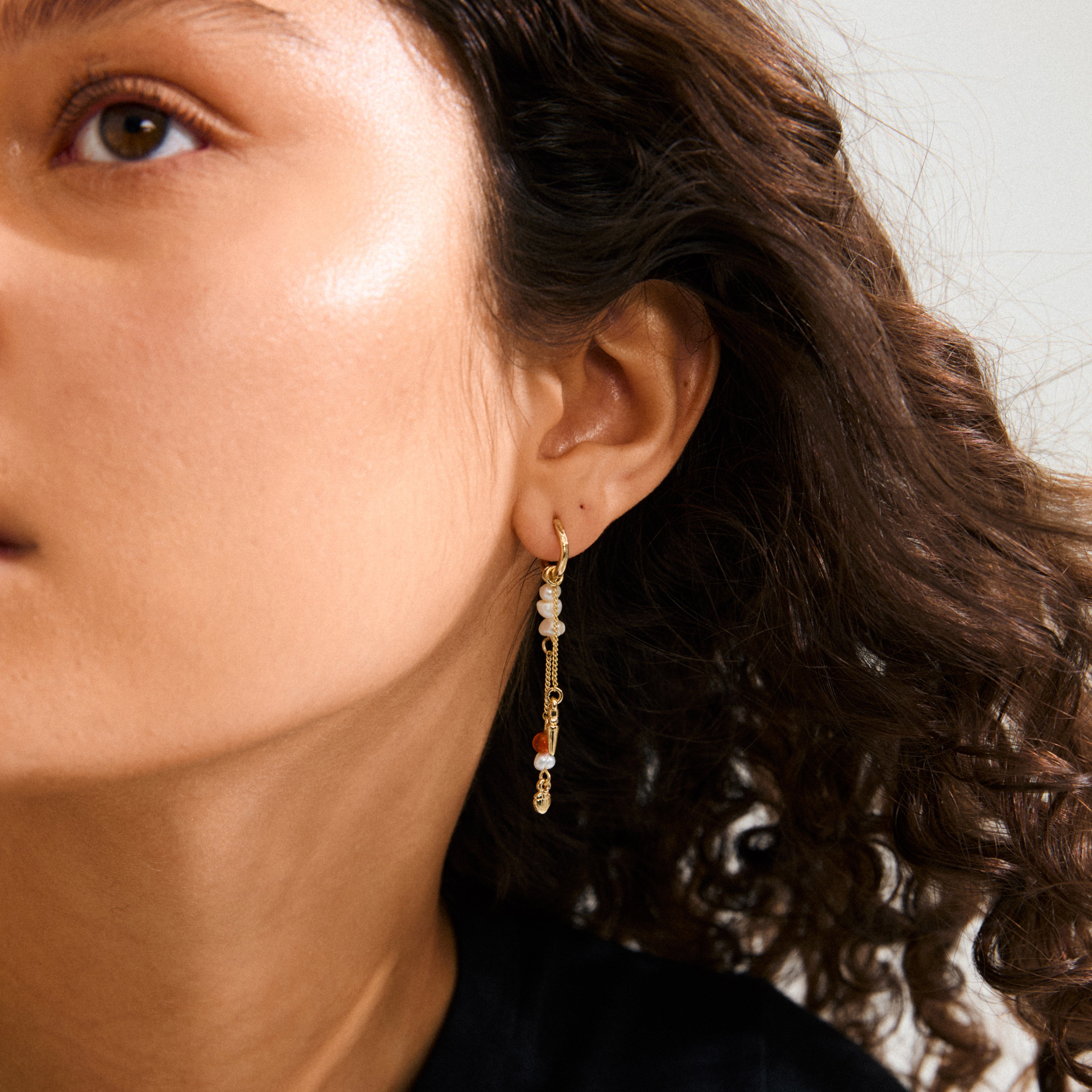 Close-up of a woman's ear wearing Pilgrim's BETWEEN gold-plated chain earrings with asymmetrical design and vibrant pendants, showcasing a bohemian and artistic style.