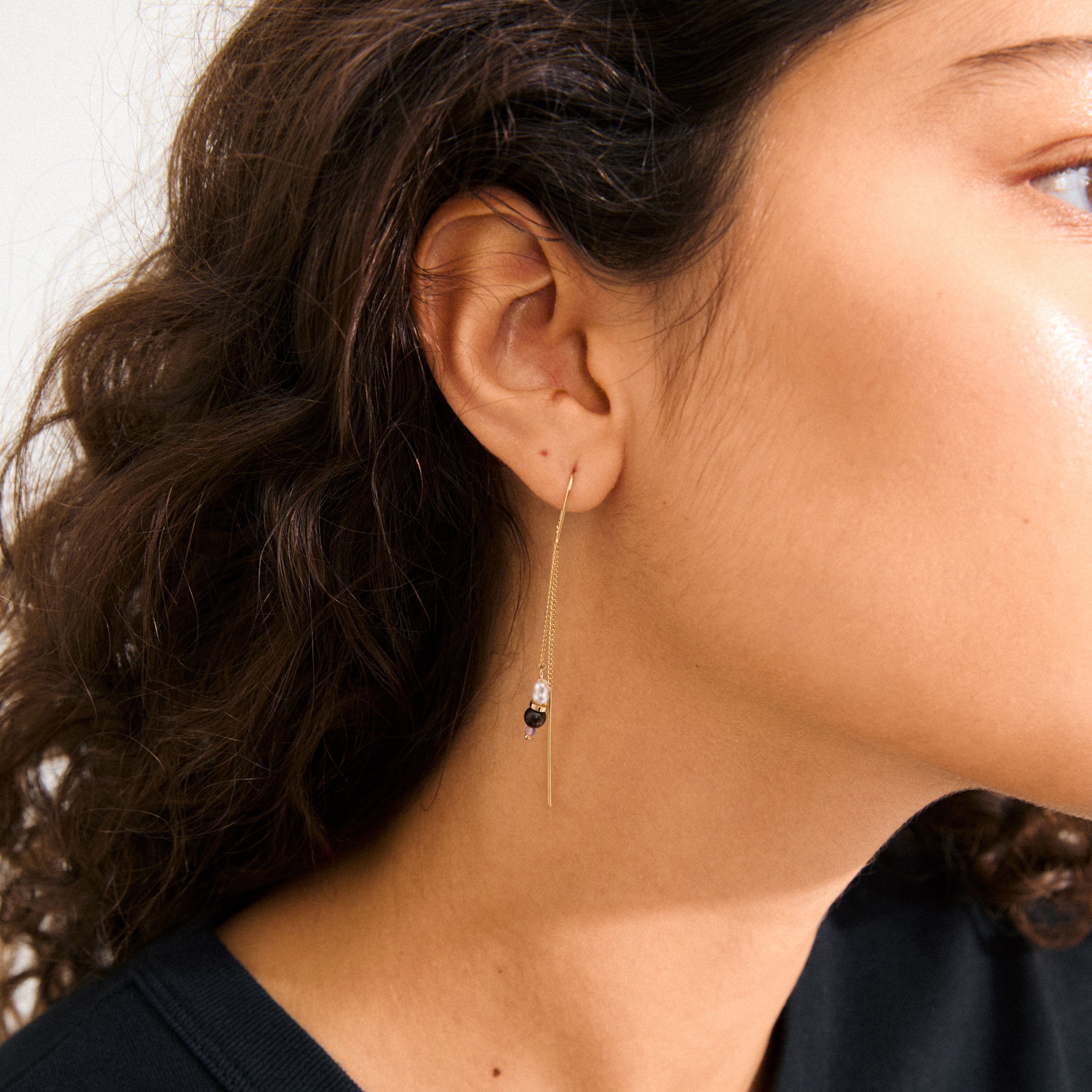 Close-up of a woman's ear showcasing Pilgrim's BETWEEN gold-plated chain earrings with asymmetrical hoops and vibrant gemstone pendants, exuding a bohemian elegance.