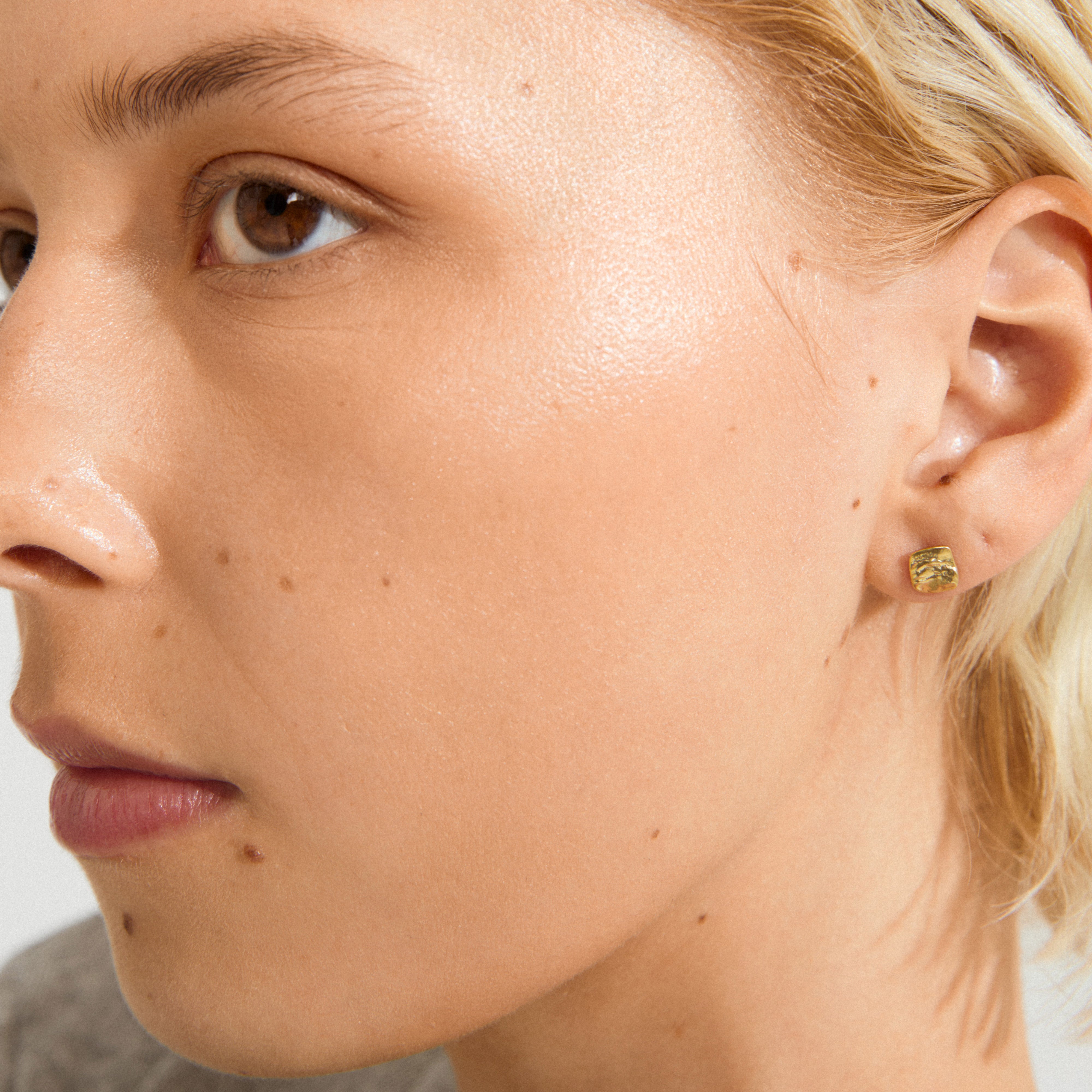 Close-up of a person wearing Pilgrim's SENSE gold-plated stud earring, featuring a small square with a hammered texture, highlighting its subtle shine and rustic elegance.