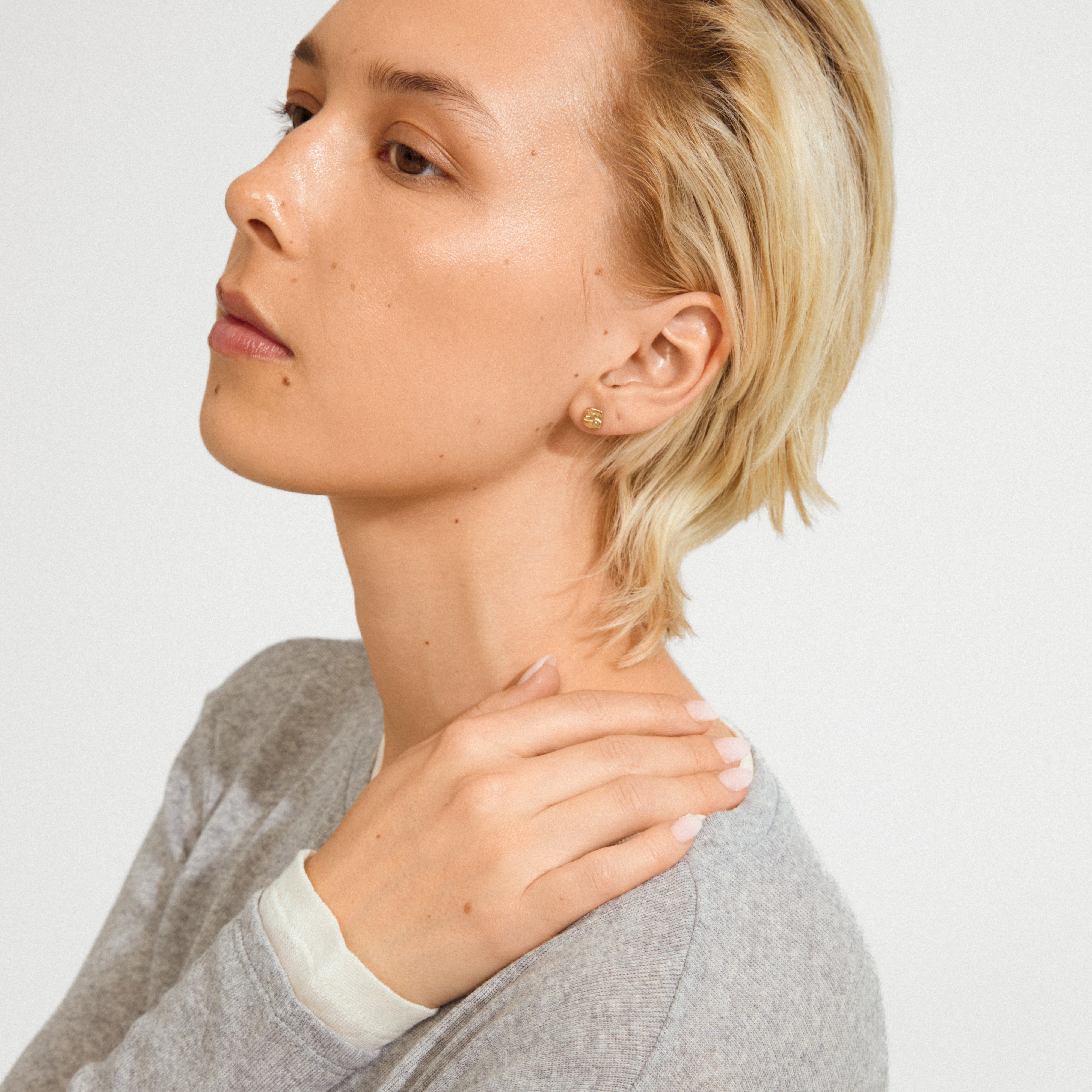 SENSE stud earrings gold-plated adorn the ear, showcasing a rustic, wavy texture that catches light, worn by a young adult in a light gray sweater.