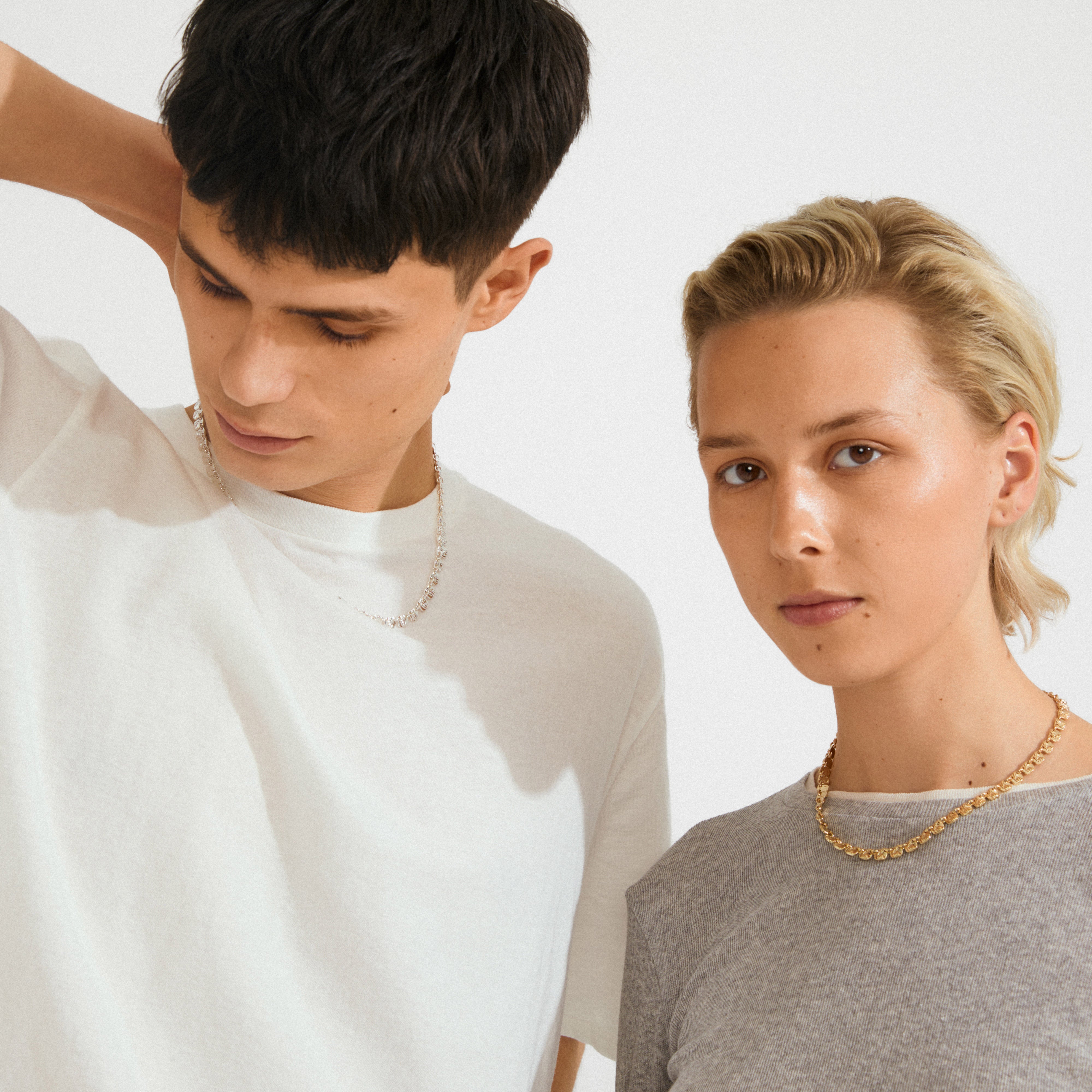 Two young adults showcasing the SENSE necklace gold-plated from Pilgrim; a stylish, gold-tone chain with small square coins, worn by the person on the right.