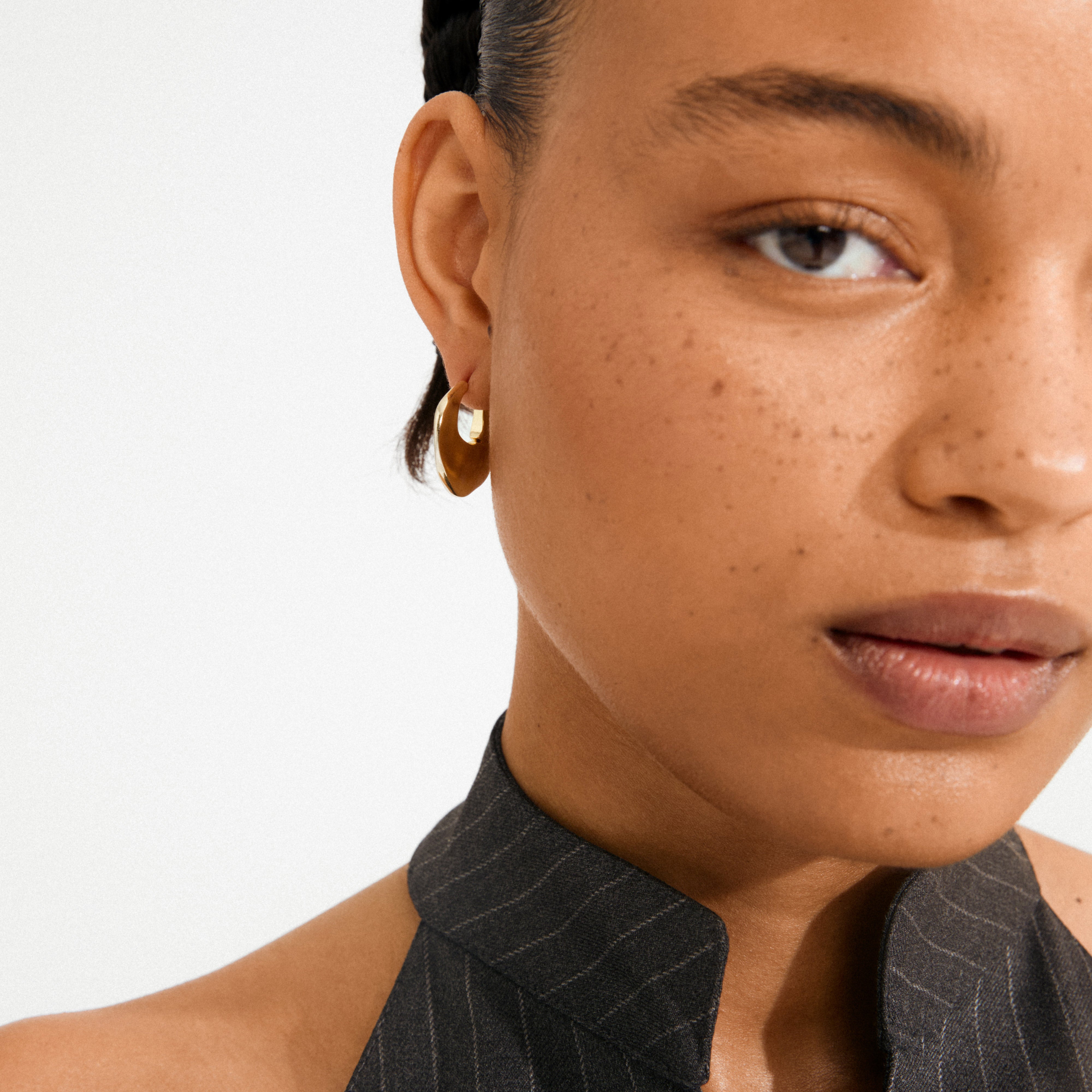 Close-up of a gold-plated SENSE hoop earring by Pilgrim on a person with freckles, glossy lips, and wearing a dark pinstripe top.