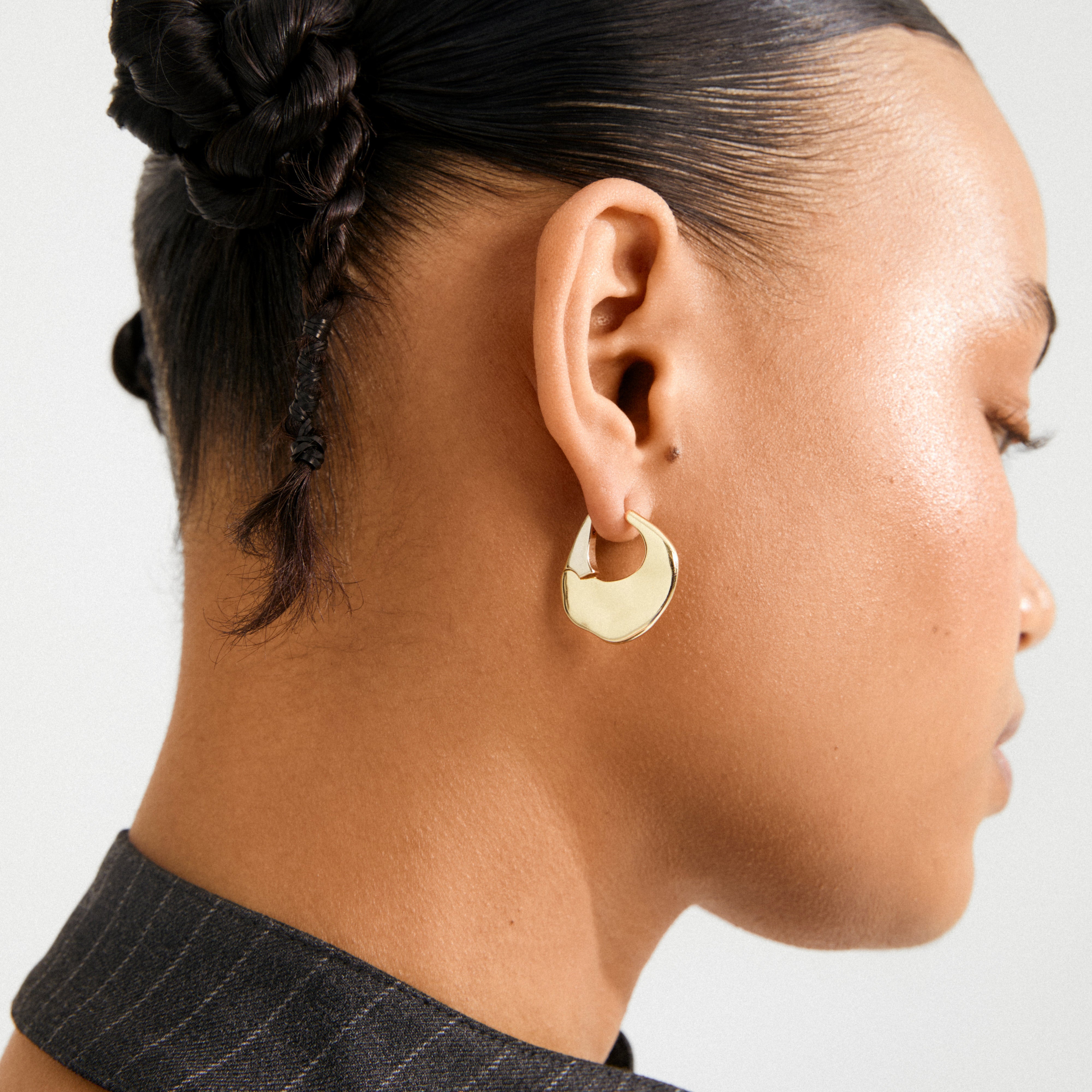 Side profile of a person wearing Pilgrim's gold-plated SENSE hoop earring, featuring an organic shape and wavy surface, styled with braided updo.