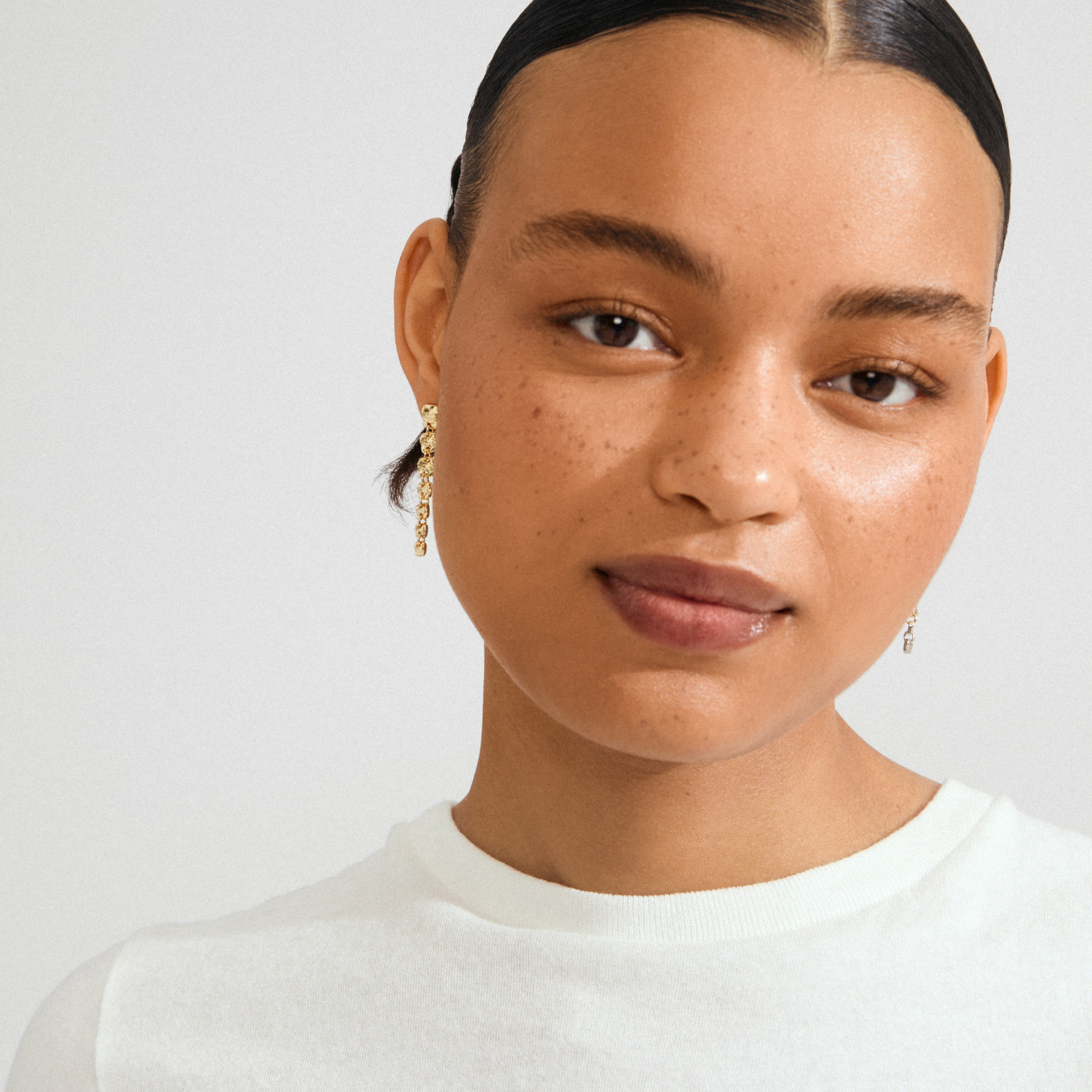 Young person wearing Pilgrim's gold-plated SENSE earrings, featuring movable chain with square coins, showcasing modern elegance with a calm expression and sleek style.