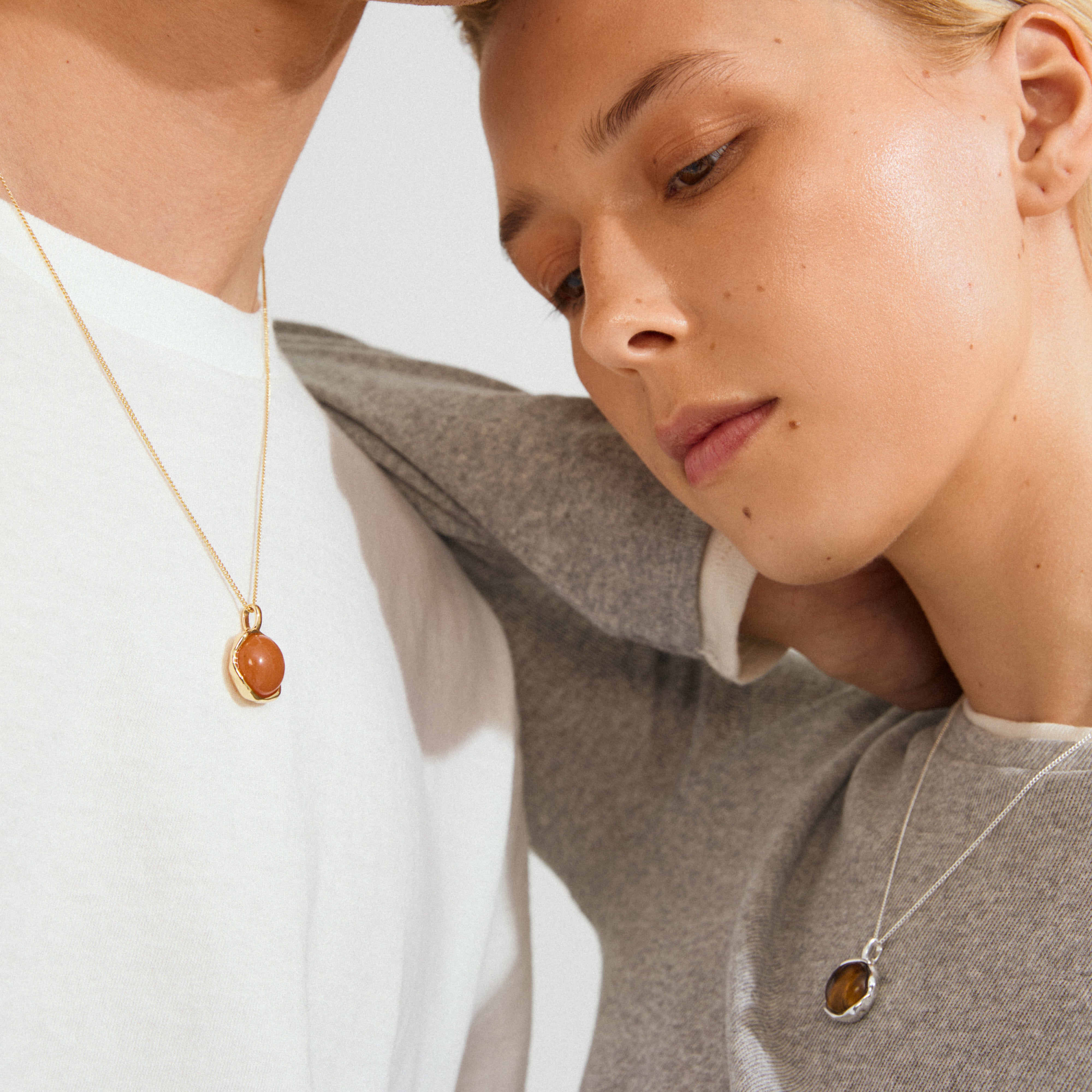 Two people wearing SENSE necklaces by Pilgrim: a gold-plated chain with red agate on white, and a silver chain with brownish gem on gray.
