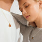 Two people wearing SENSE necklaces by Pilgrim: a gold-plated chain with red agate on white, and a silver chain with brownish gem on gray.