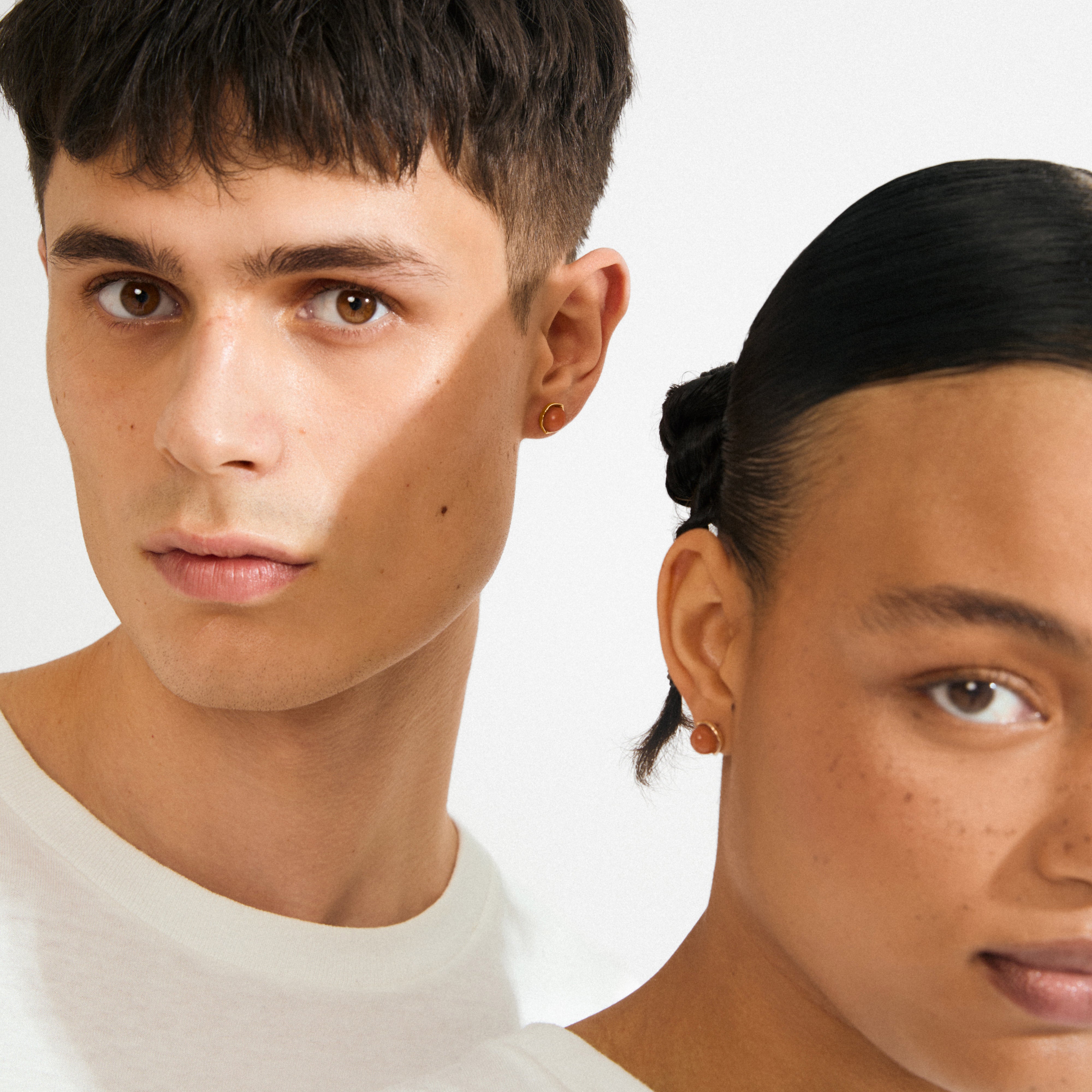 Two models wear Pilgrim's SENSE earrings, gold-plated with Red Agate stones, showcasing minimalist elegance in a close-up portrait.