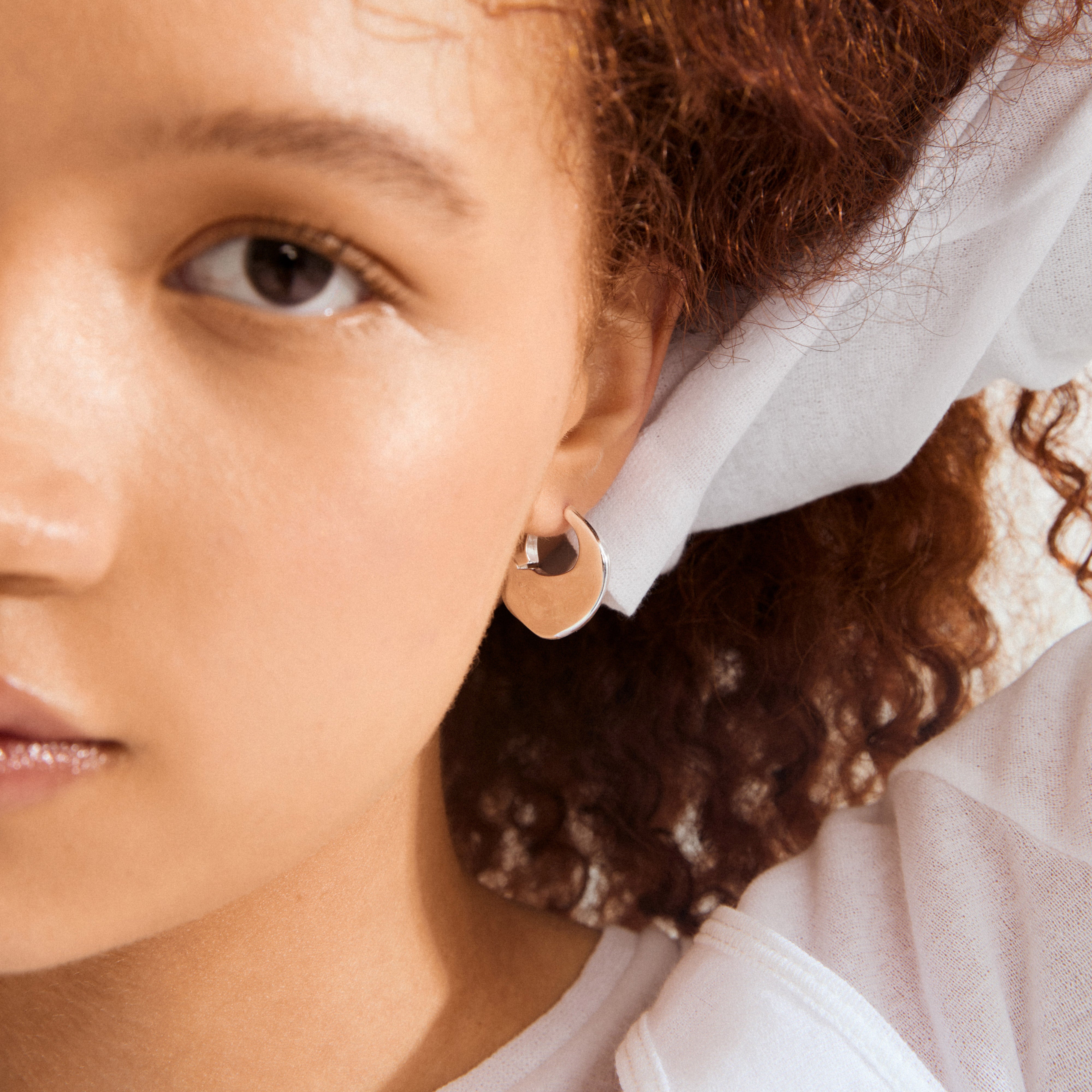 Close-up of SENSE silver-plated hoop earring on left ear, featuring a polished, wavy design. Partially visible reddish-brown curls under a white headscarf.