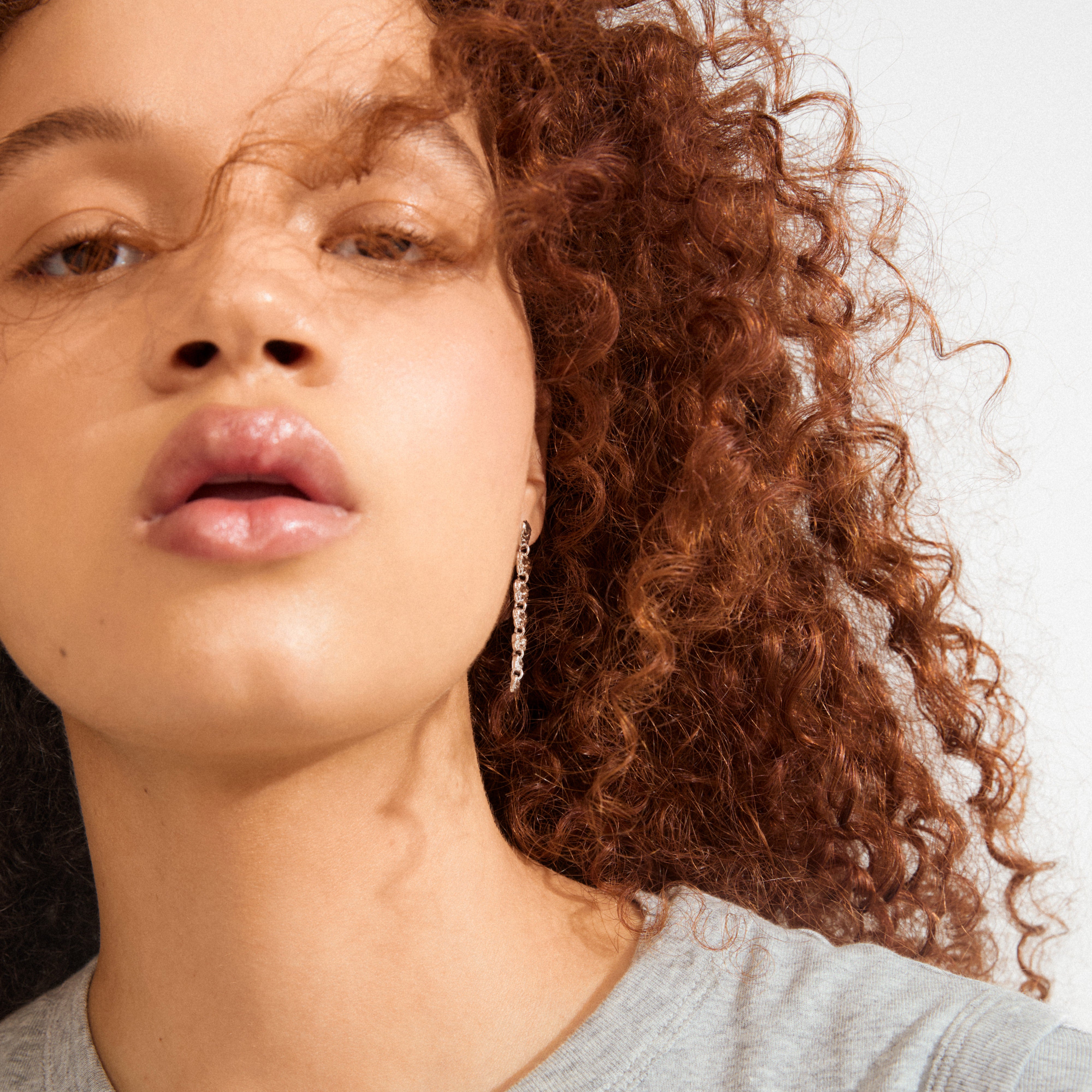 Close-up of SENSE earrings silver-plated by Pilgrim, featuring a dangling silver earring on a person with glossy lips and long, curly reddish-brown hair.