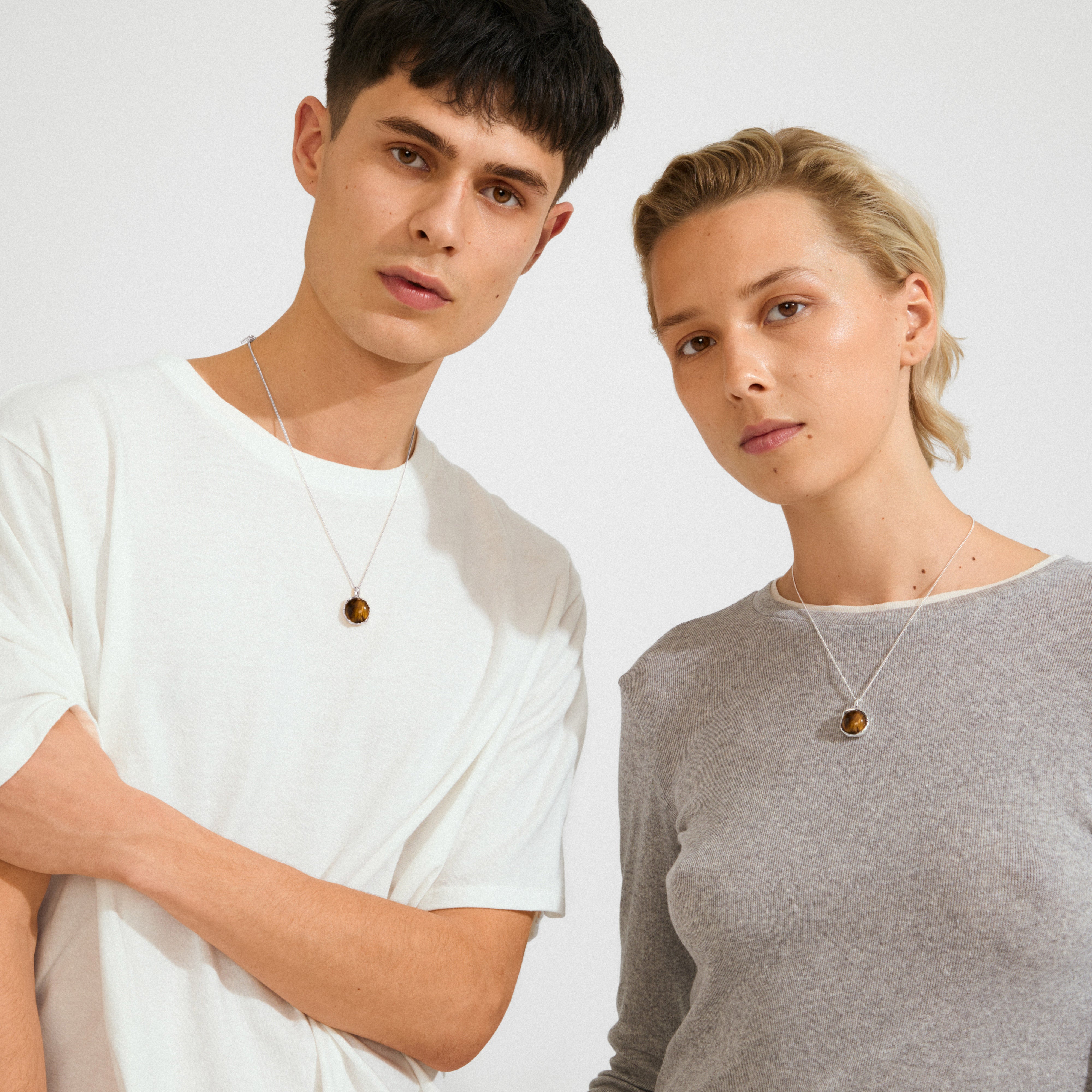 Two young adults wearing Pilgrim's SENSE necklace, featuring a silver chain and a round Tiger Eye stone pendant, symbolizing strength and confidence.