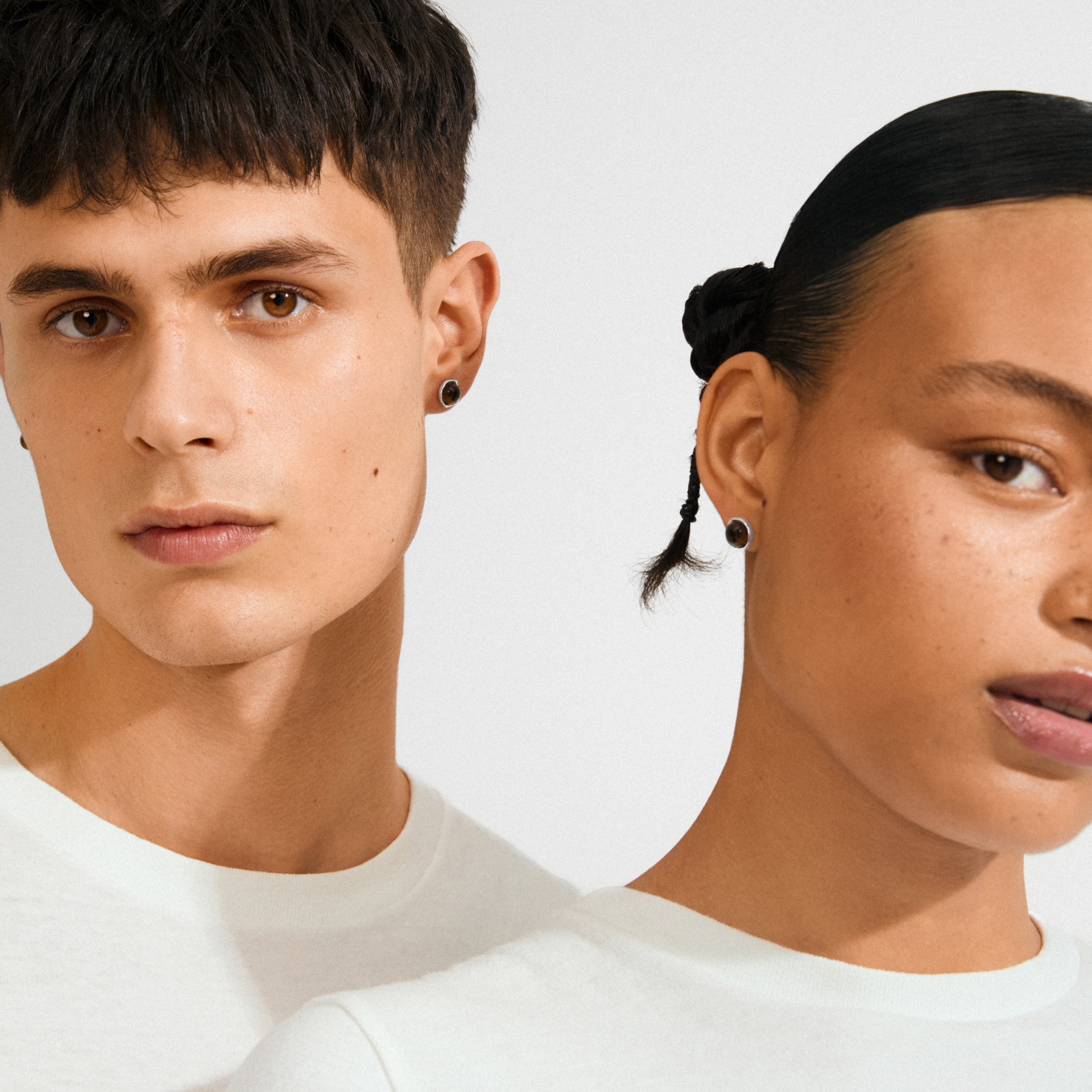 Two young adults wearing SENSE earrings by Pilgrim, showcasing silver-plated settings with Tiger Eye stones, symbolizing strength and balance, against a simple backdrop.
