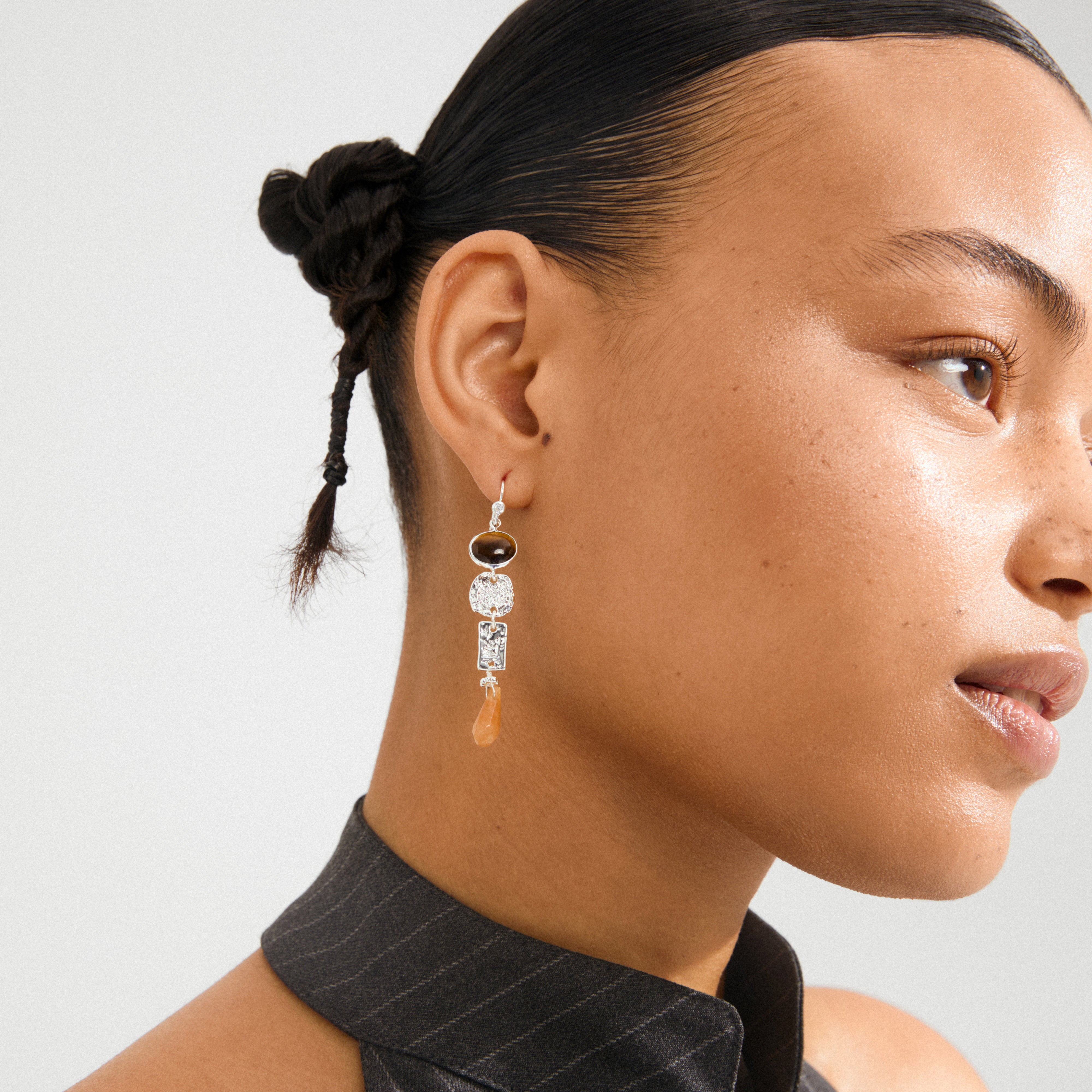 Woman wearing SENSE earrings, silver-plated with dark stone, ornate silver links, and teardrop bead, styled with braided buns and pinstripe halter top.