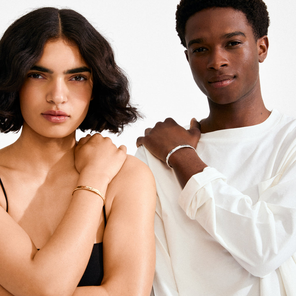 Two people showcase Pilgrim's EMBER bracelet gold-plated; left person highlights its wavy, textured design with a black top; right person sports a silver-tone bracelet.