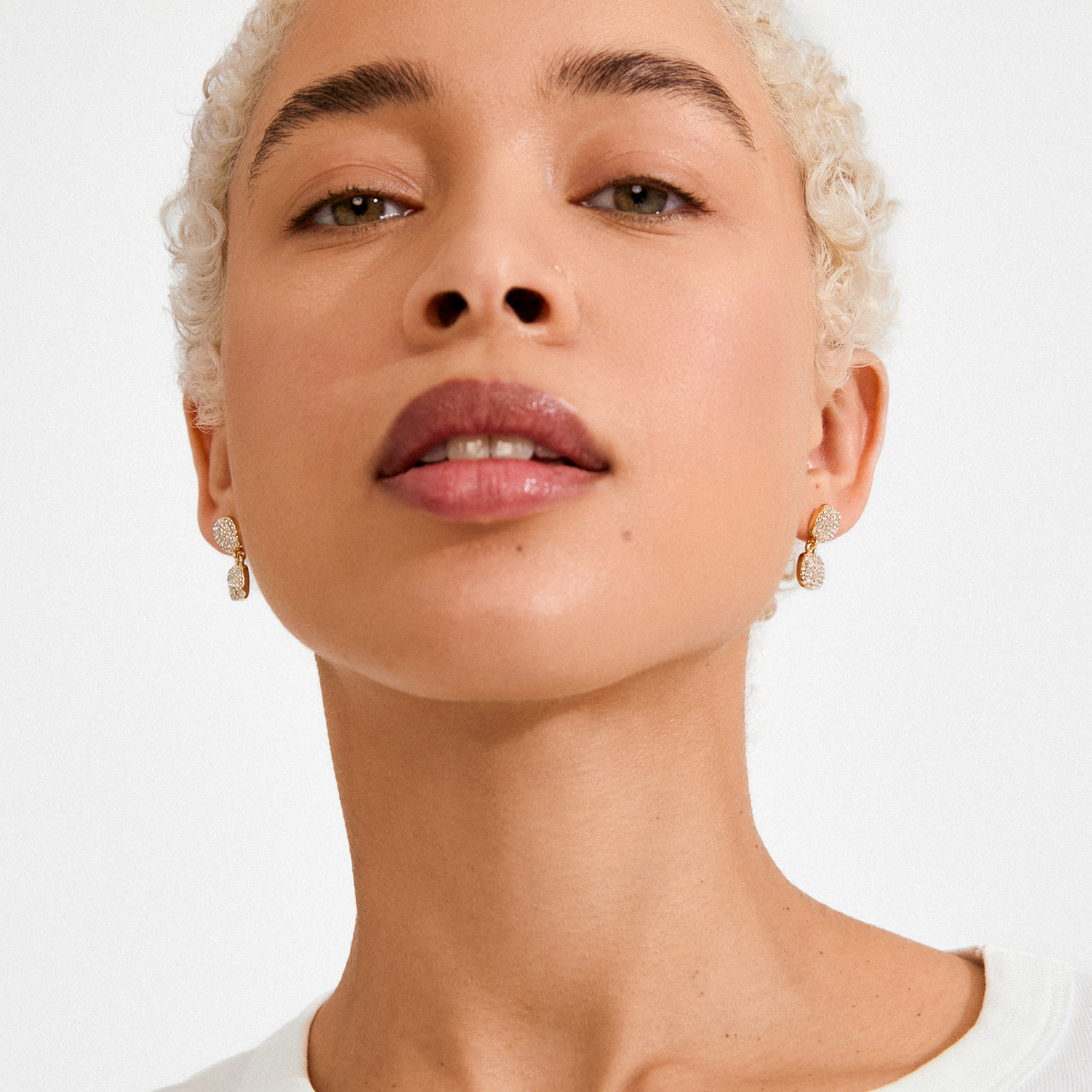 Close-up of a person wearing EMBER earrings gold-plated, showcasing small, gold-tone drop earrings encrusted with sparkling Preciosa crystals, highlighting their elegance and sophistication.