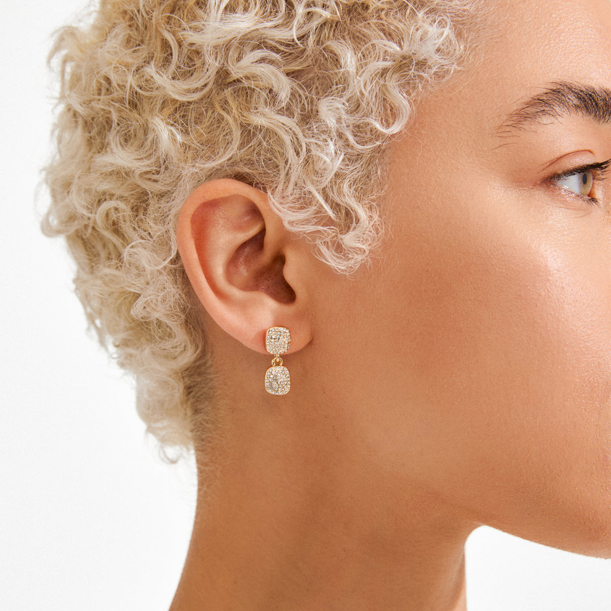 Close-up of a person's ear wearing Pilgrim's EMBER gold-plated earrings with two sparkling, square pavé-set crystal clusters, showcasing Scandinavian-inspired elegance and sustainability.