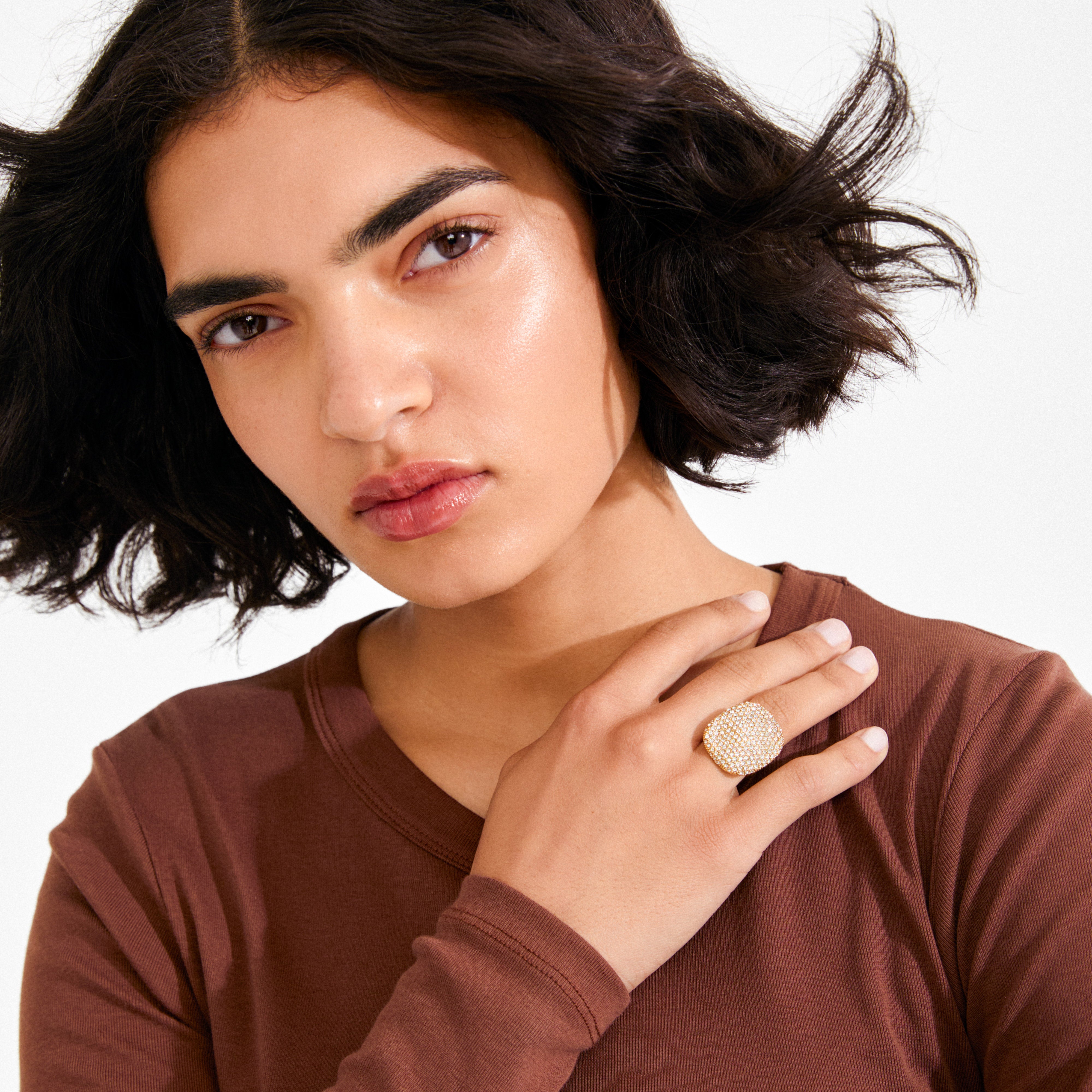 Person wearing a long-sleeved brown top showcasing the EMBER ring gold-plated by Pilgrim, featuring a bold, wavy square centerpiece with sparkling crystals on their ring finger.