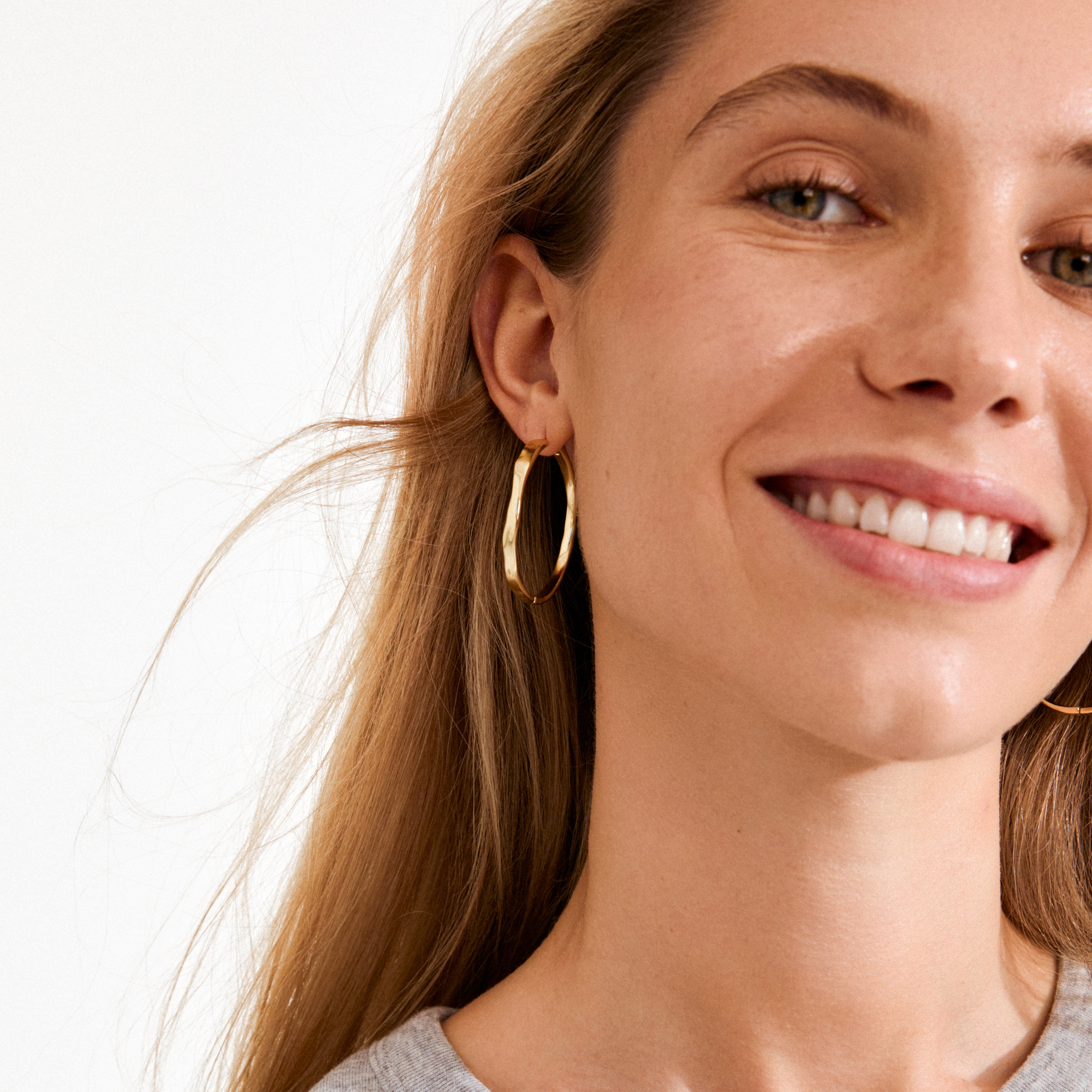 Close-up of a smiling woman wearing gold-plated EMBER hoop earrings from Pilgrim, showcasing their subtle wavy design and texture.