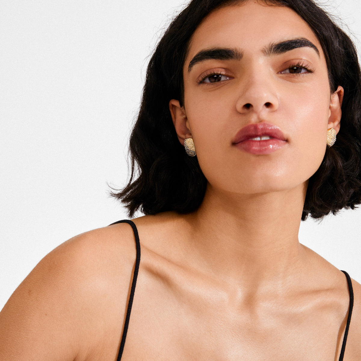 Young woman wearing gold-plated EMBER earrings by Pilgrim, featuring a bold, textured design with sparkling crystals, complementing her dewy skin and glossy lips.