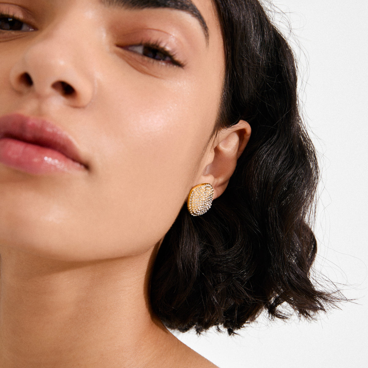 Close-up of a woman's ear wearing Pilgrim's gold-plated EMBER earrings, featuring a bold, irregular shape with sparkling Preciosa crystals for a chic, luxe touch.