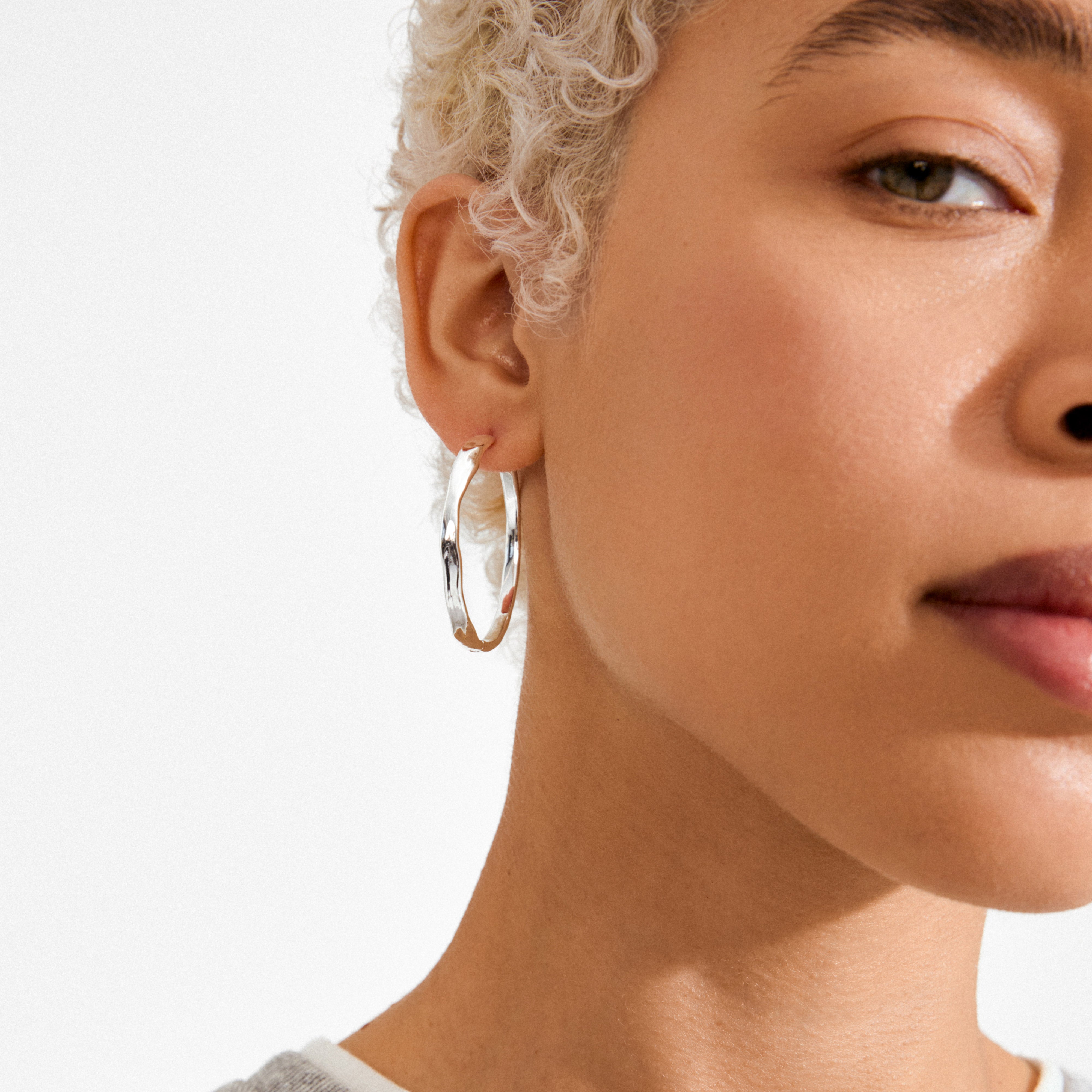 Close-up of a person's ear wearing EMBER hoop earrings, silver-plated with a subtle wavy design, highlighting Pilgrim's blend of timeless and trendy style.